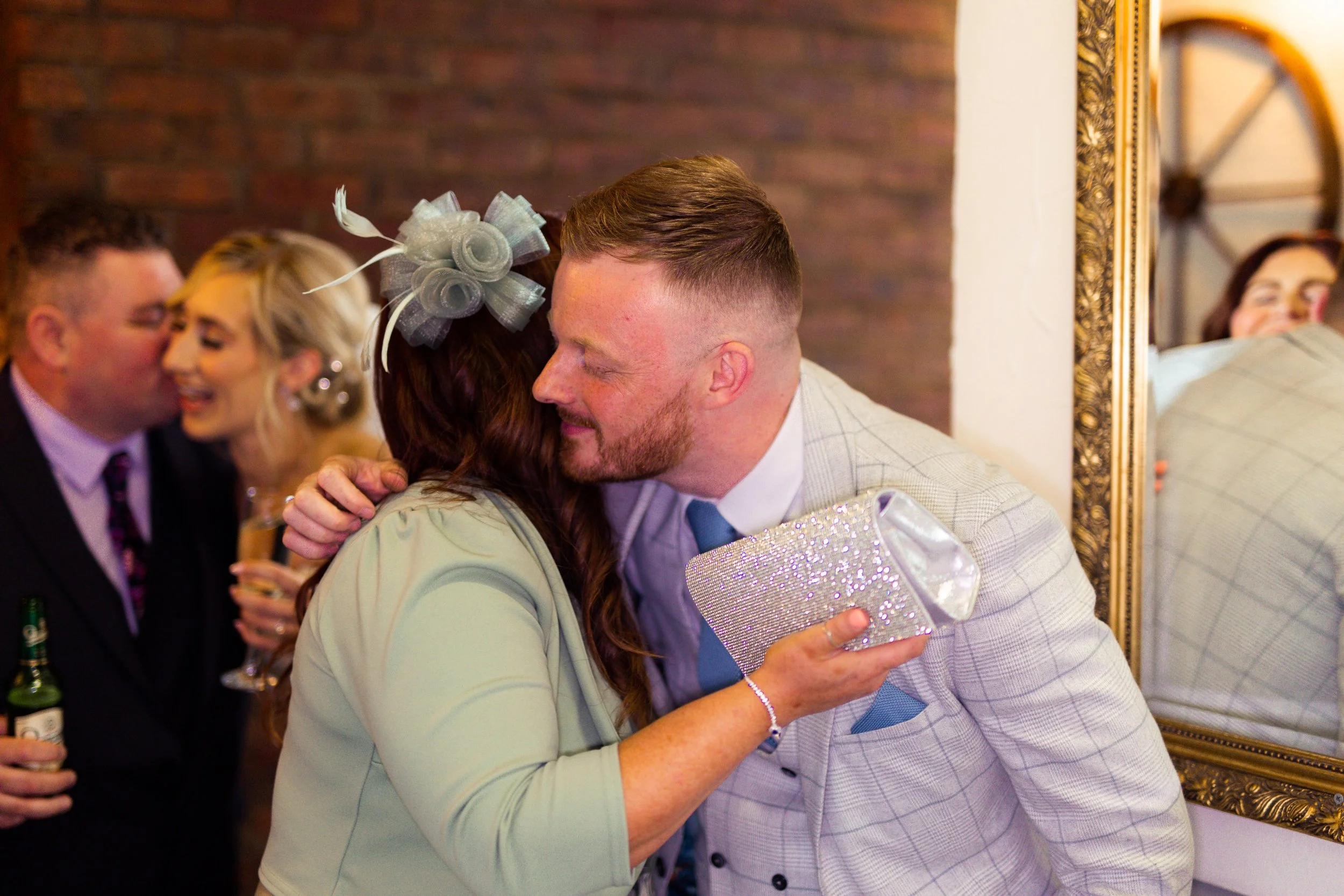 A woman hugging a man at a social gathering, with other people in the background, including a man and woman who are kissing, and a woman lying on a bed reflected in a mirror.