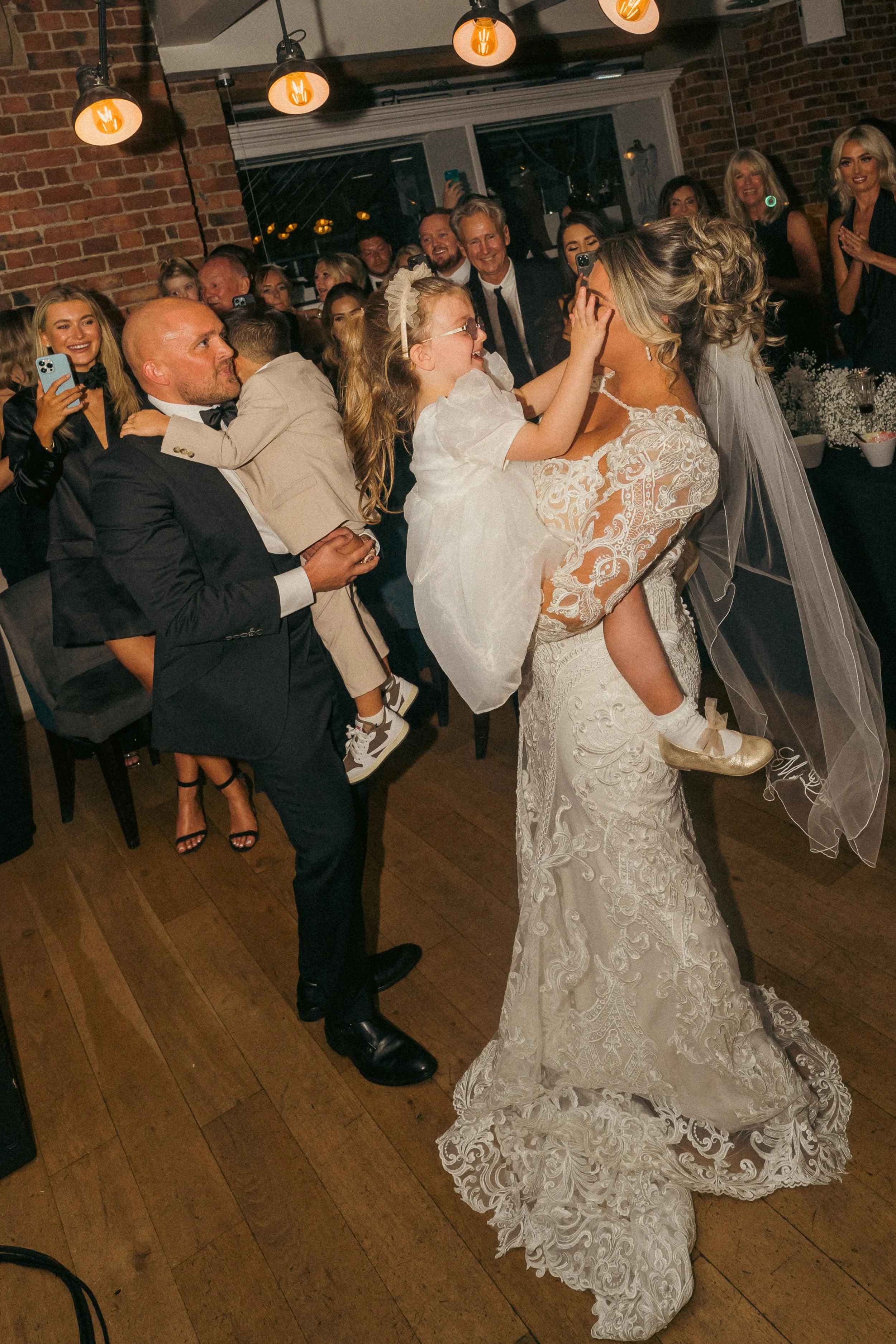 A bride in a white lace wedding gown holds a young girl wearing a white dress, smiling and touching the bride's face, while surrounded by wedding guests in formal attire at an indoor reception.