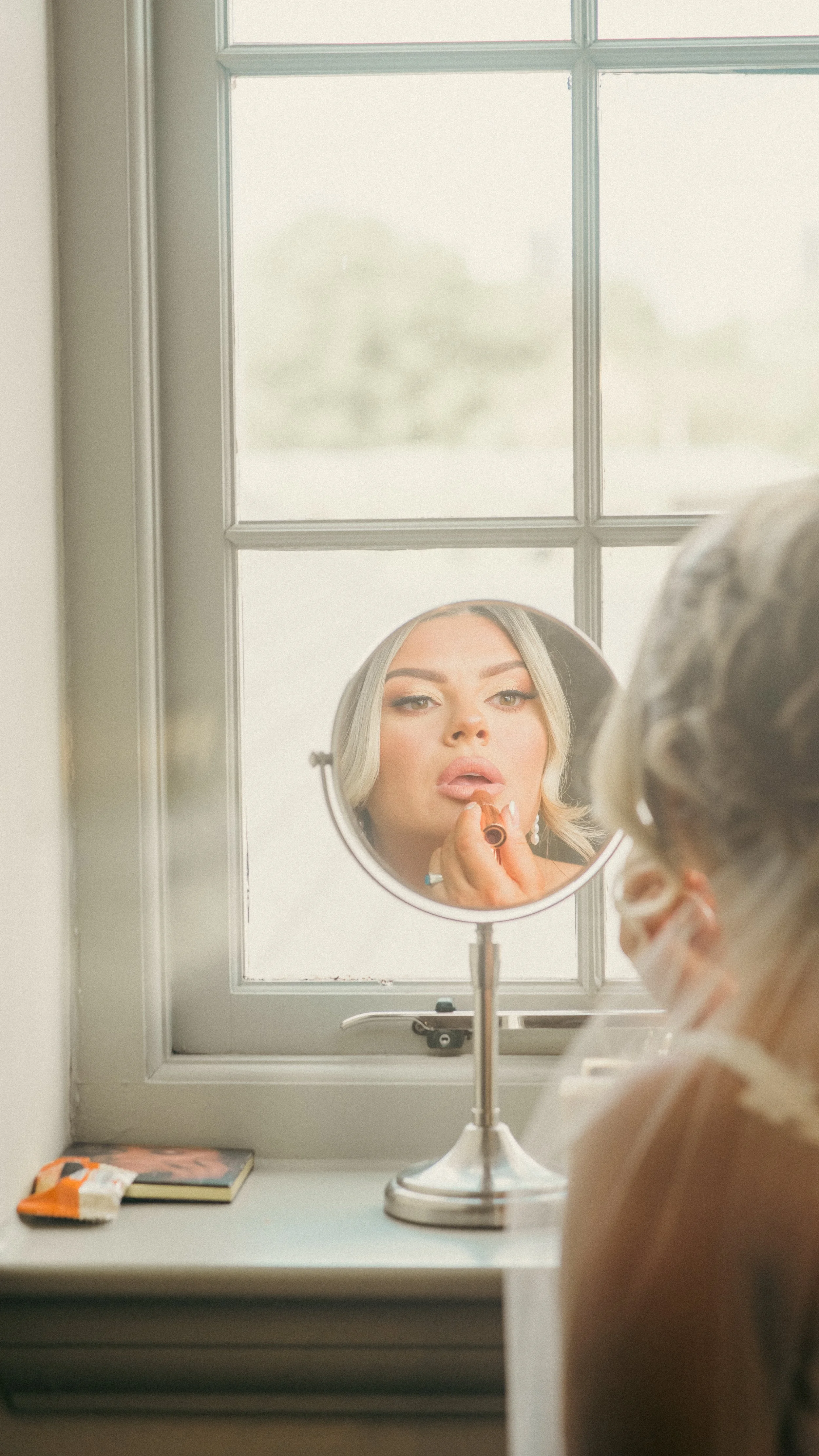 A woman is applying lipstick while looking in a round mirror on a windowsill. She is reflected in the mirror, and the window behind her shows a blurry outdoor scene.