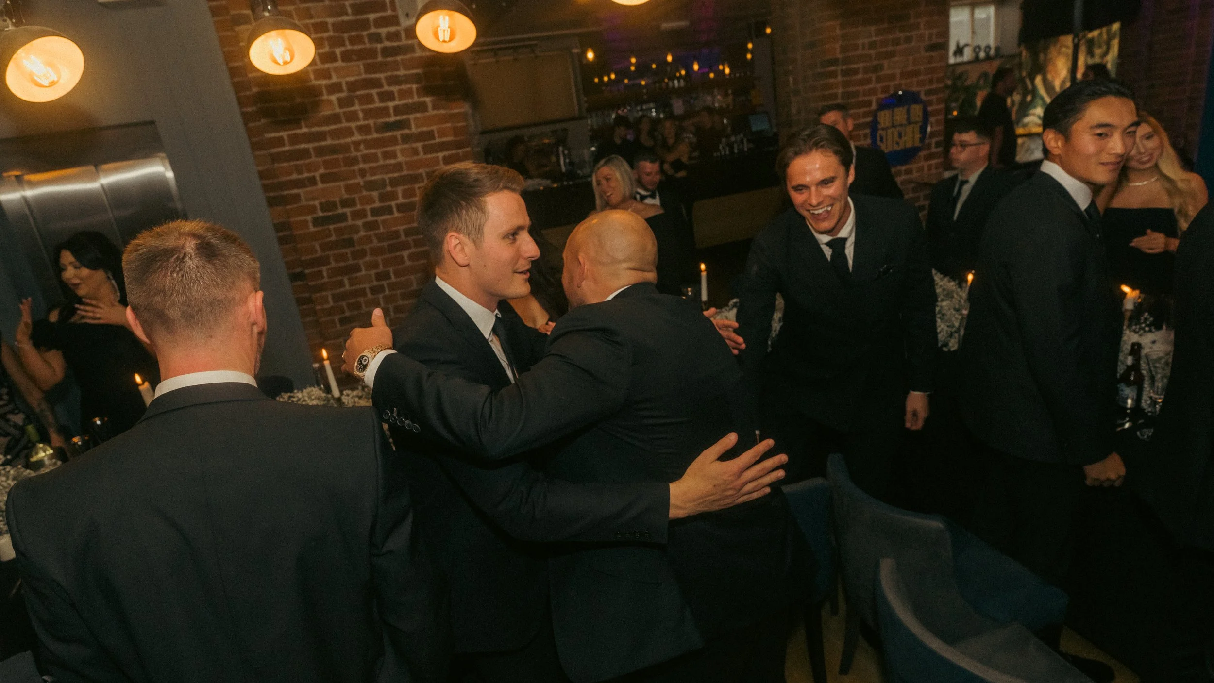 People dressed in formal black suits and dresses at a social event or celebration, hugging and smiling in a warmly lit room with brick walls and hanging lights.
