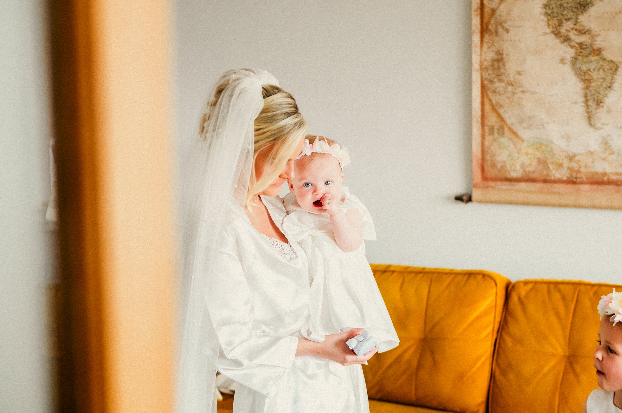 A woman dressed as a bride holding a young girl dressed in white. The woman is leaning in close to the girl, who has a floral headband and is touching her mouth with her finger. They are indoors, with a map on the wall and a yellow couch in the backg