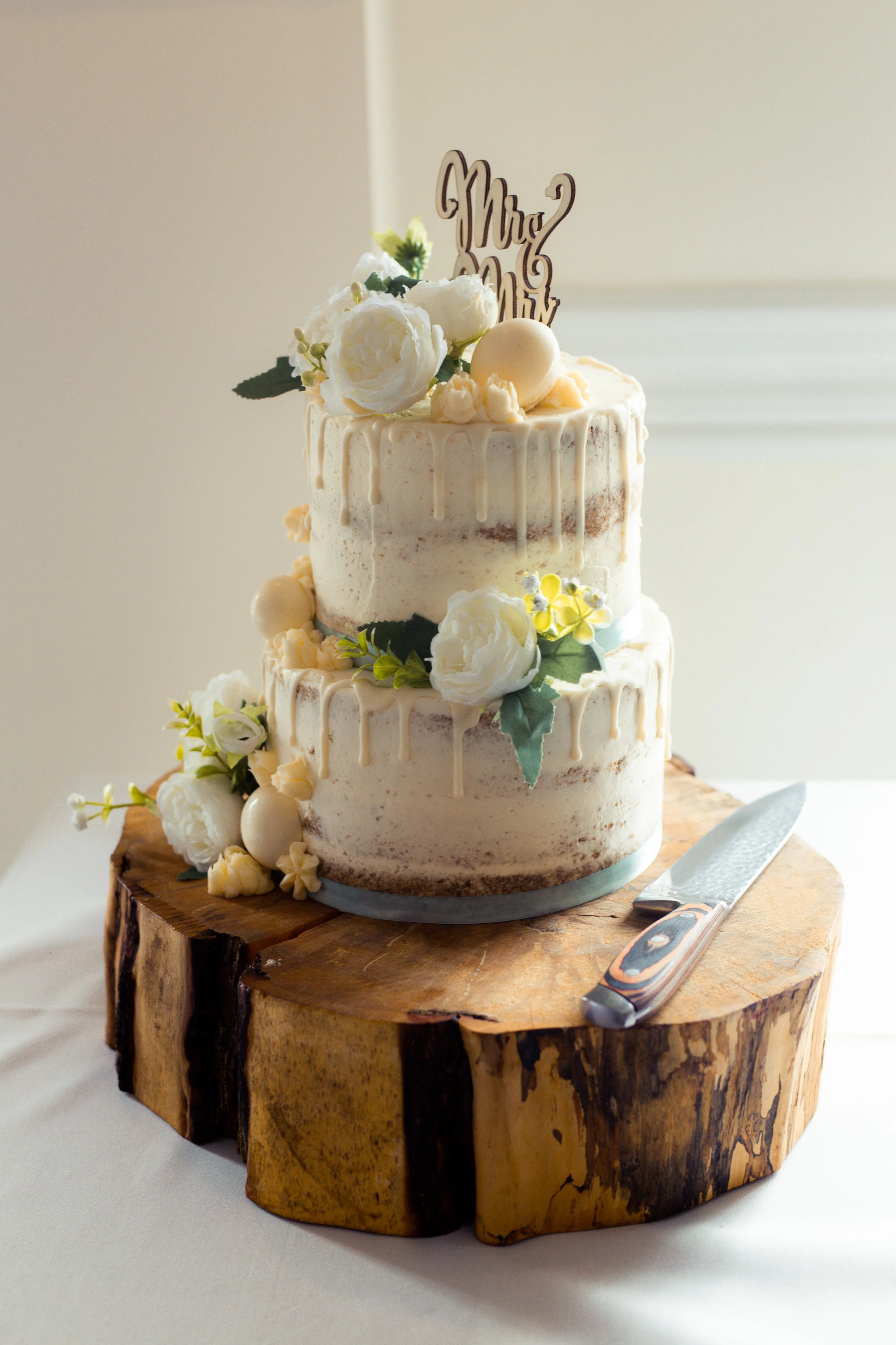 A two-tiered wedding cake decorated with white flowers, green leaves, and a topper that says "Mr. and Mrs." on a rustic wooden base, with a cake knife beside it.