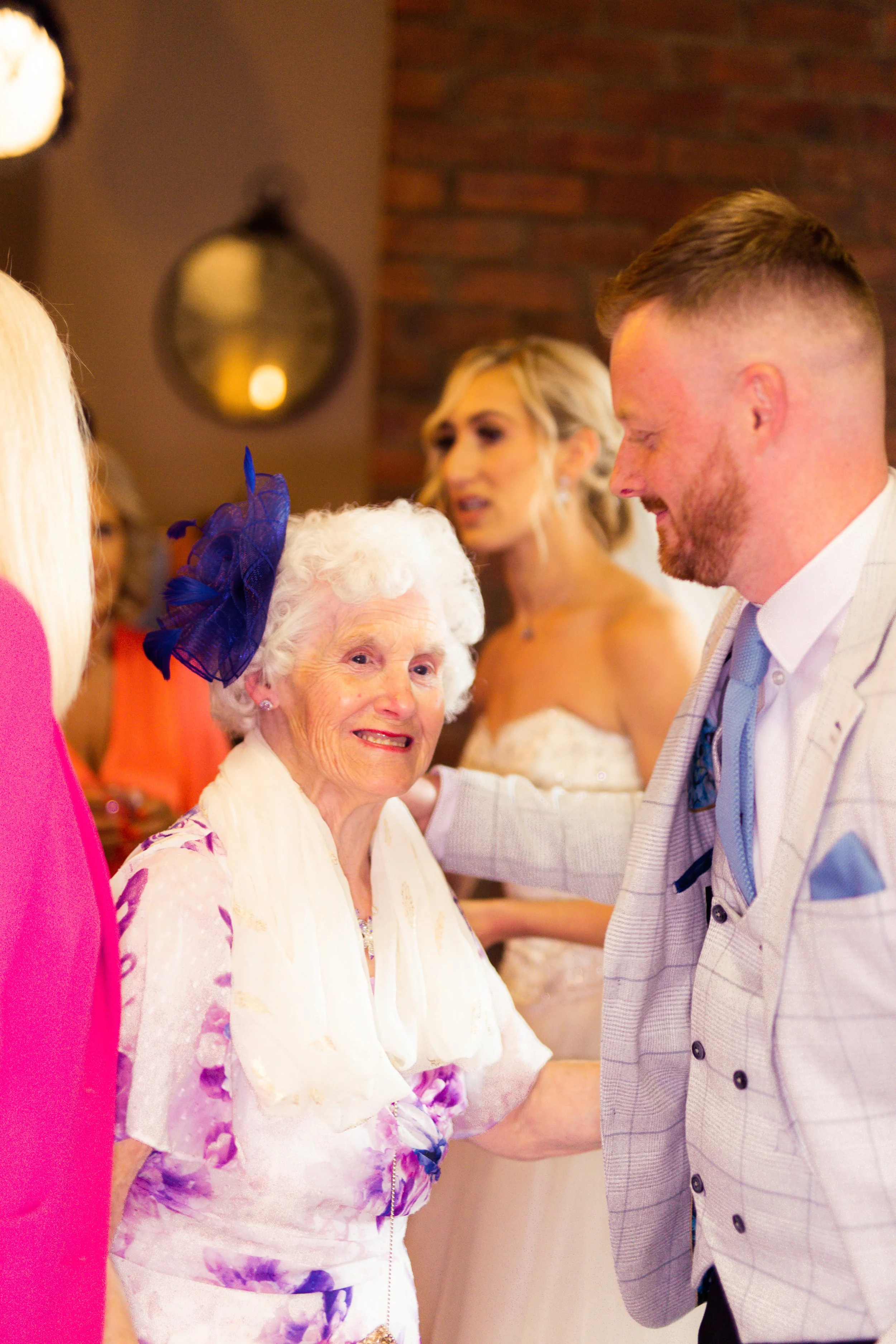 A wedding reception with a smiling elderly woman in a floral dress and purple hat talking to a man in a light-colored suit. A bride in a white wedding dress stands behind them, looking on.
