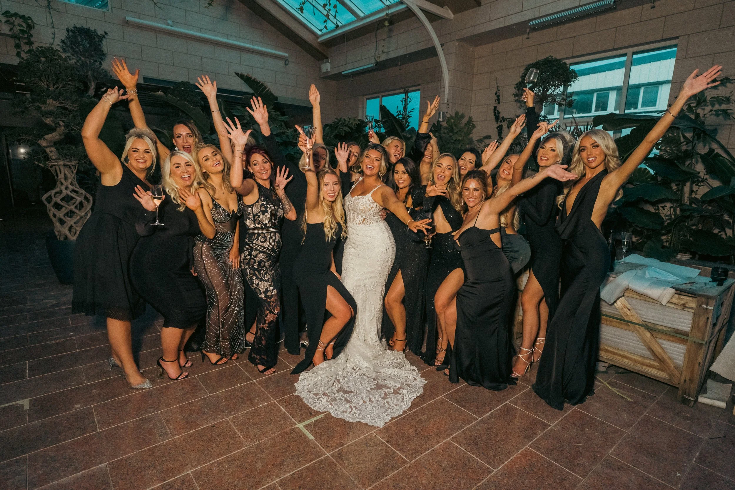 A group of women at a celebration, some in black dresses, with one in a white wedding dress, in an indoor venue with large windows and plants.