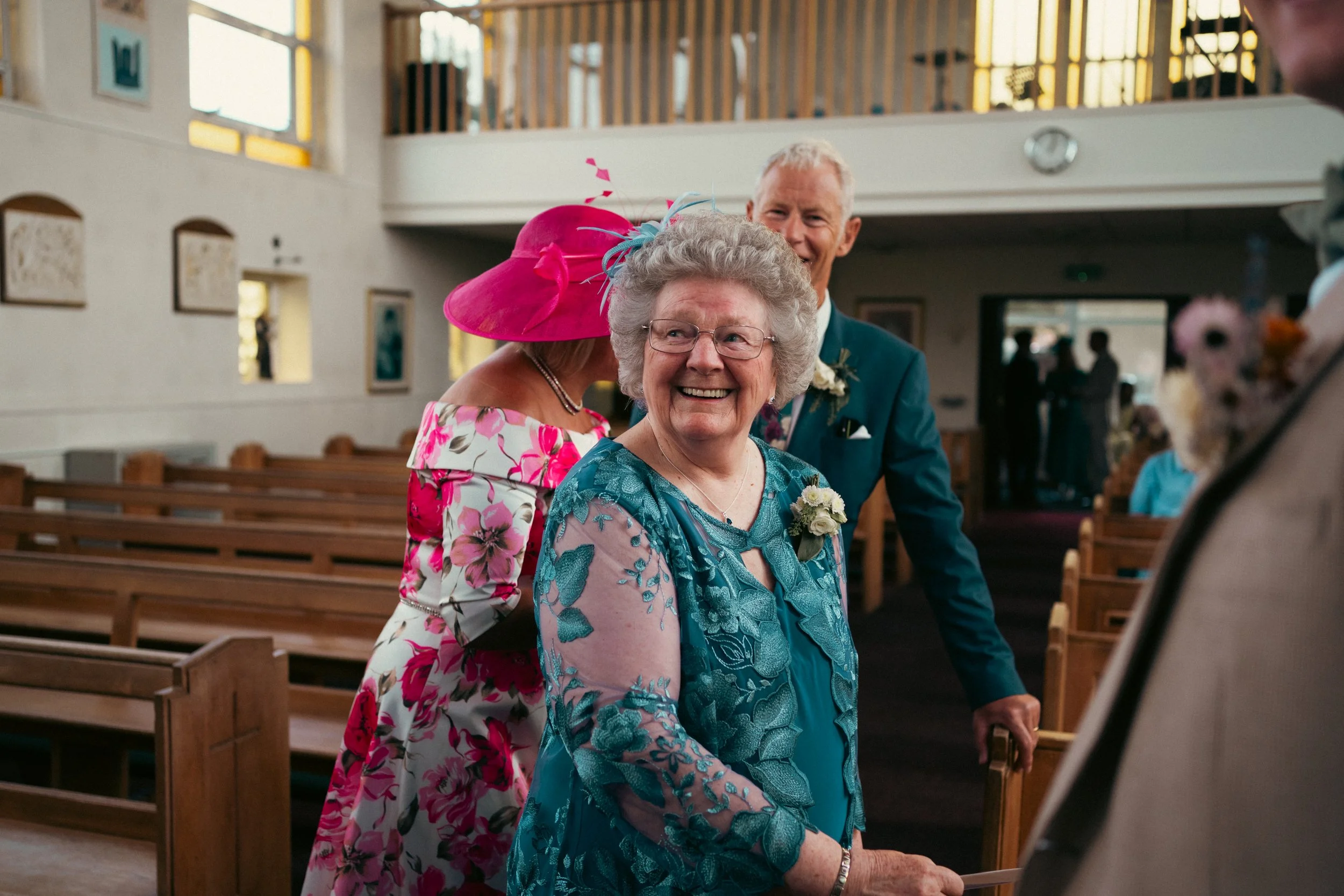 Joyful elderly woman smiling inside a church with wedding guests around her, including a man in a suit and a woman in a floral dress with a pink hat.