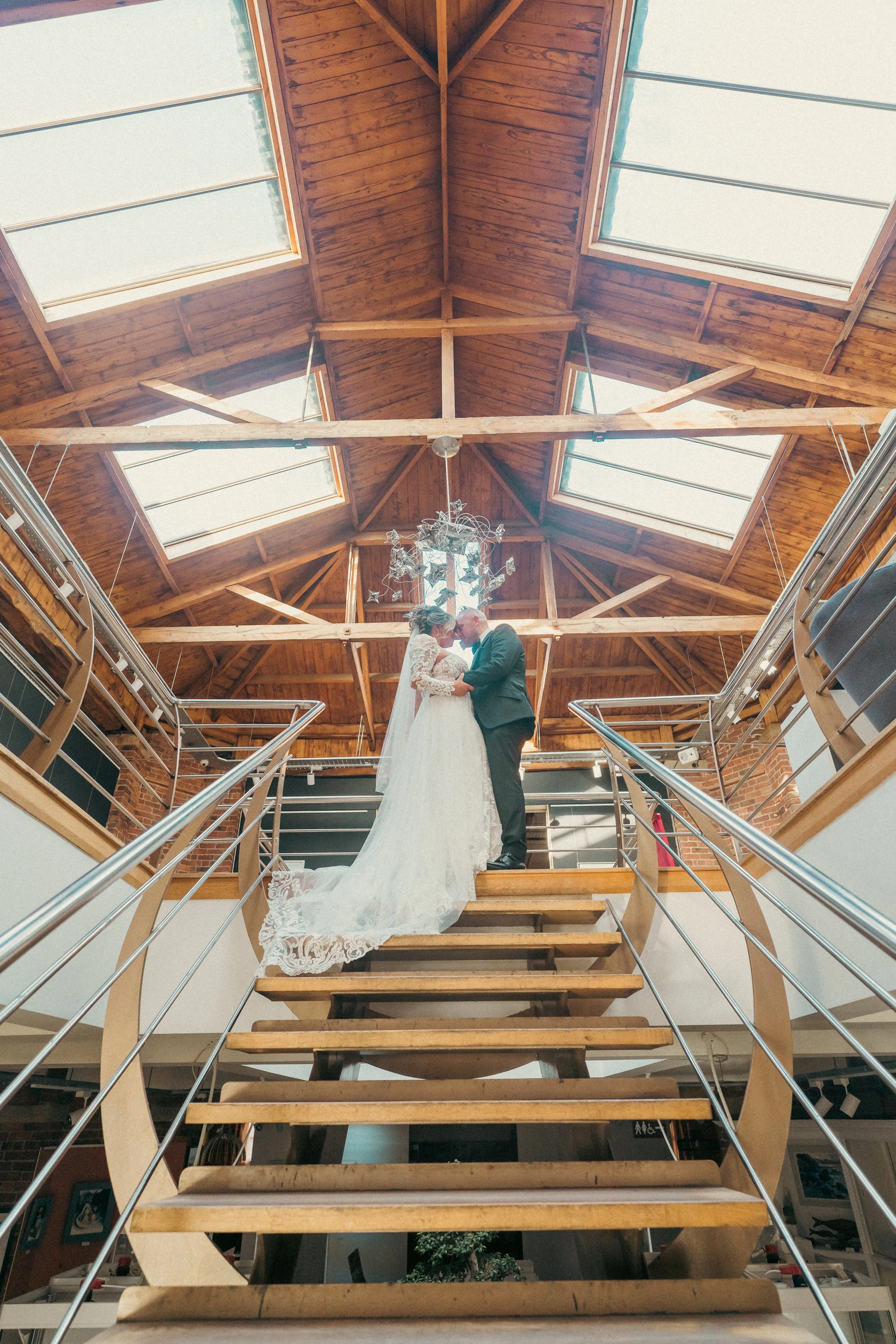 A bride and groom standing on a wooden staircase, holding hands and touching foreheads, inside a venue with a wooden ceiling and large skylights.
