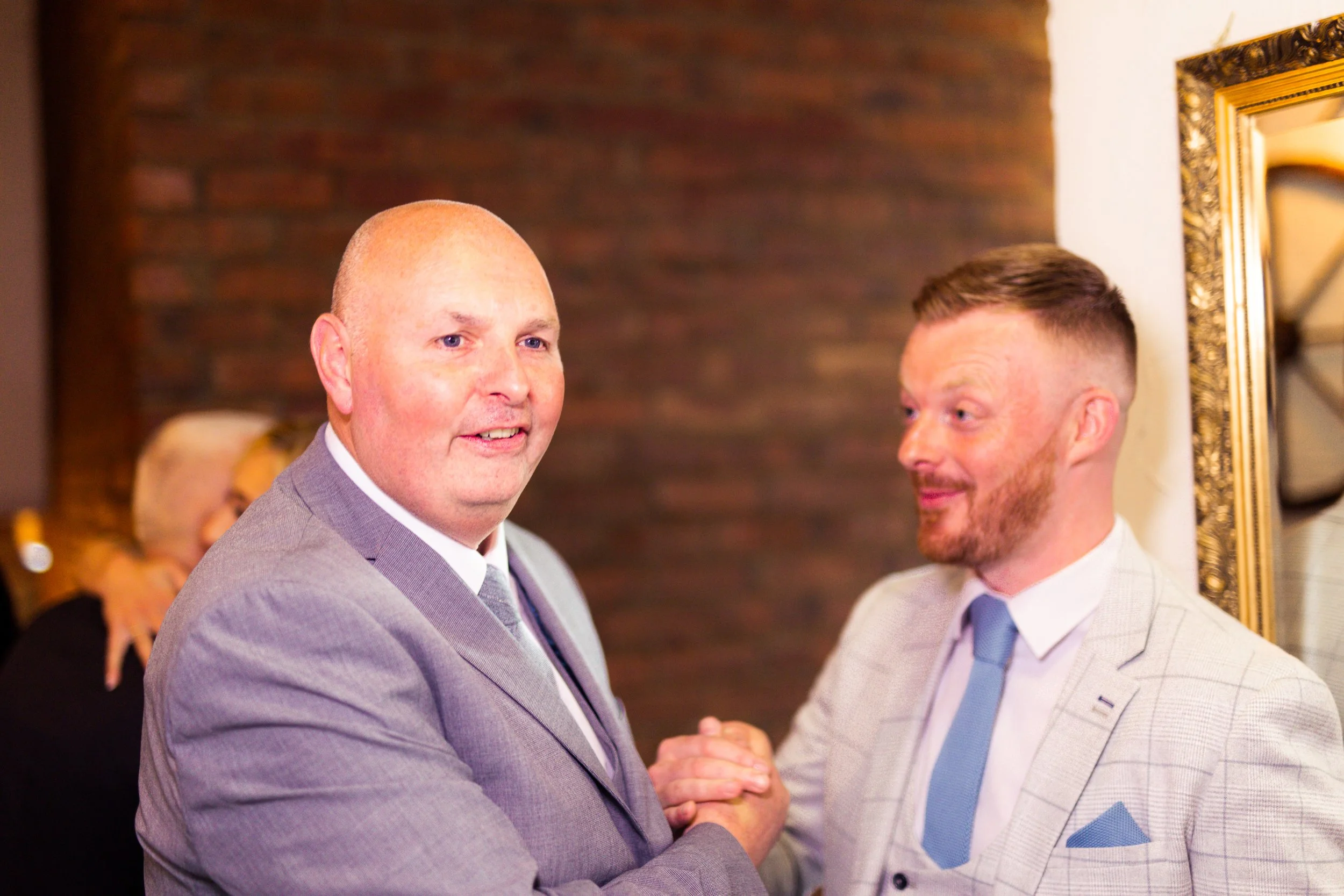 Two men shaking hands at a formal event, one with a bald head and the other with short, styled hair, both wearing light-colored suits.
