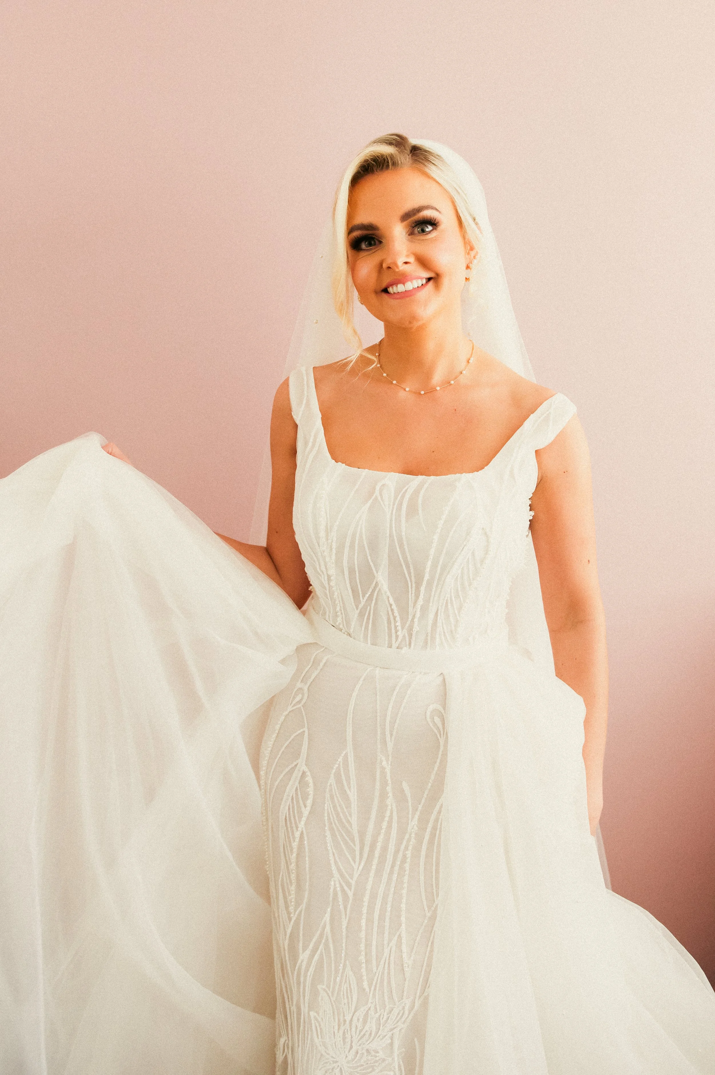 A smiling bride in a white wedding gown holding up her dress, standing against a pink wall.