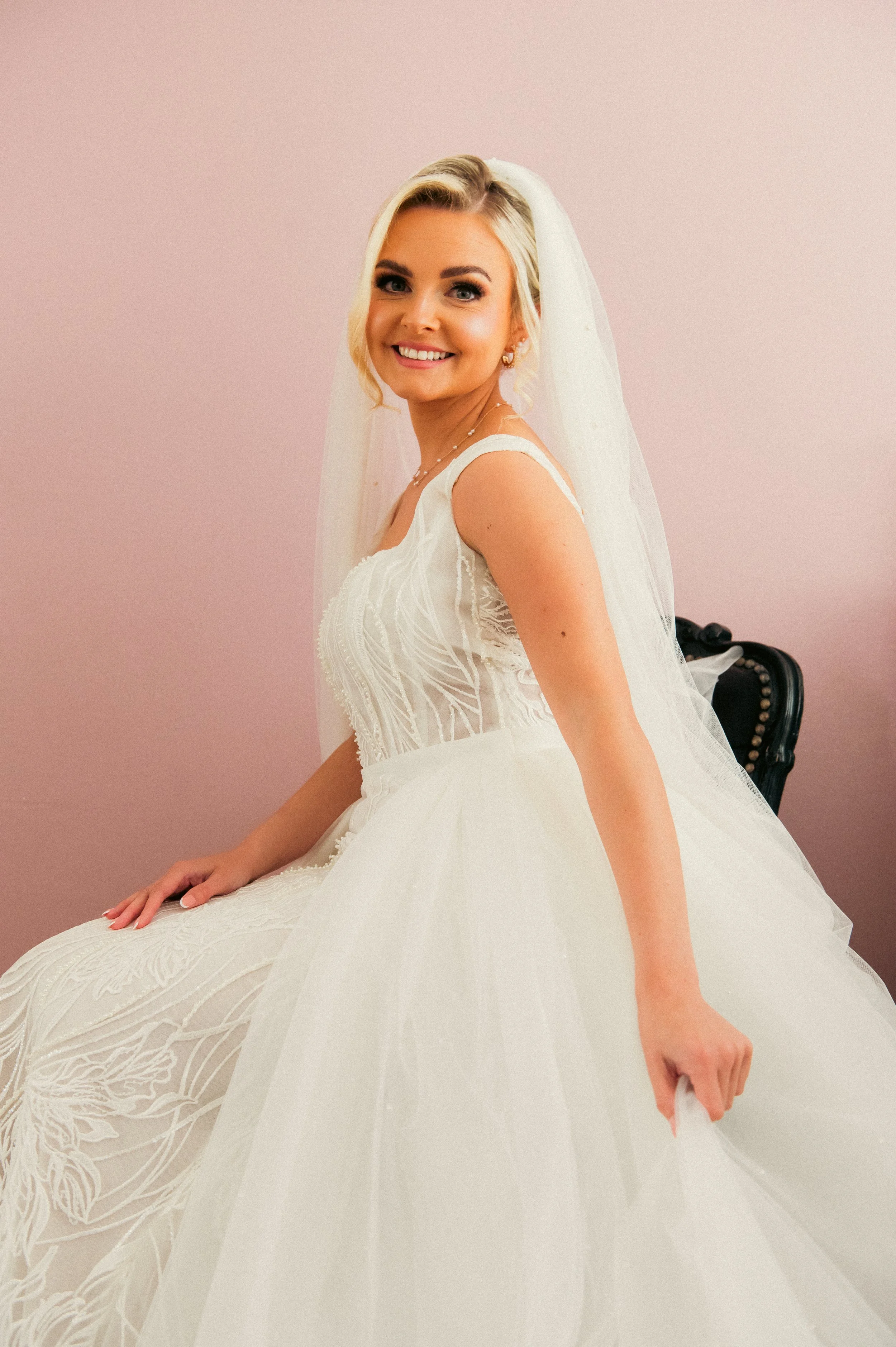 A smiling woman in a white wedding dress with a veil, sitting on a black chair against a pink wall.
