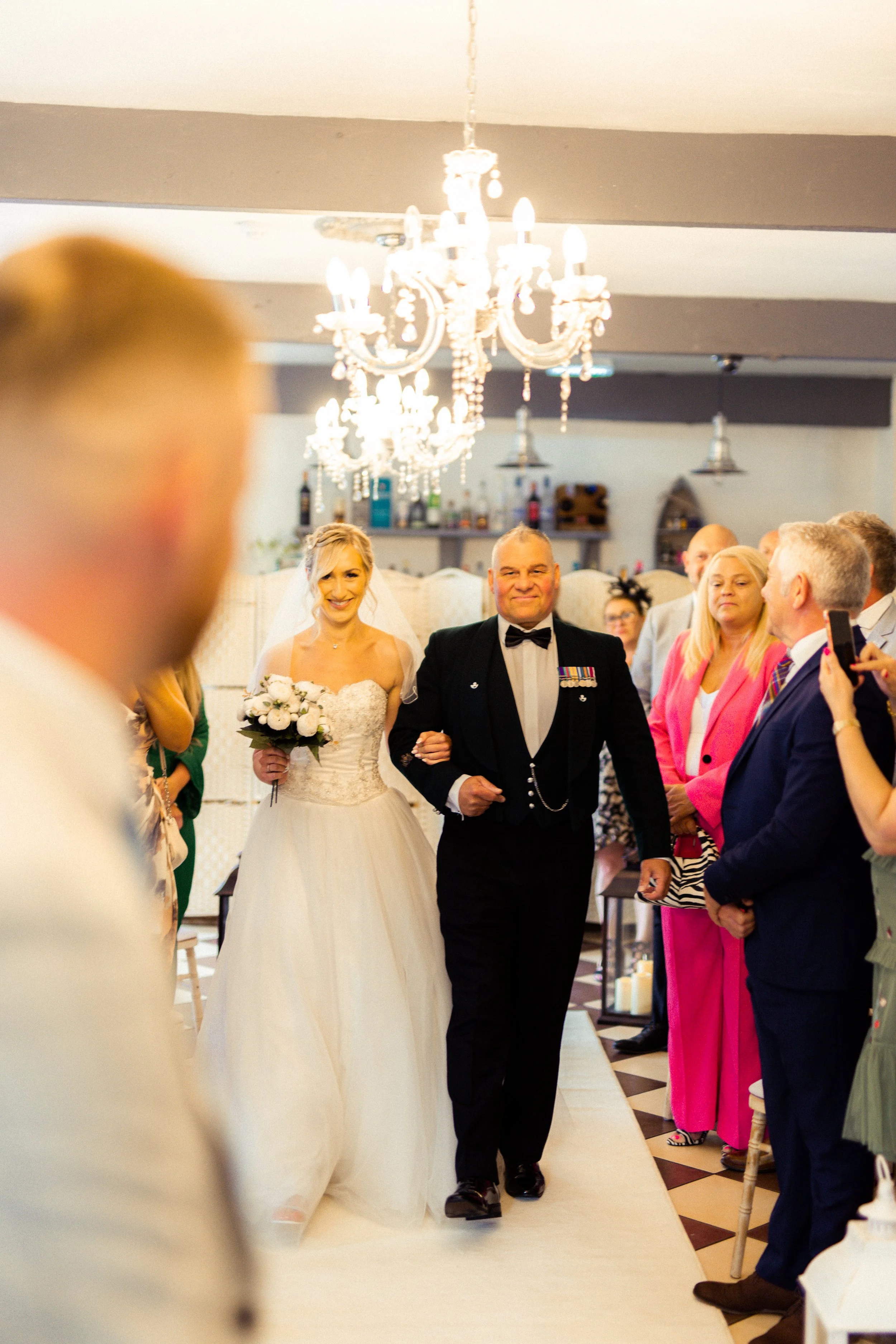 Bride walking down the aisle with a man in a military uniform at her side during a wedding ceremony. Guests are watching and taking pictures in a decorated indoor venue with chandeliers.