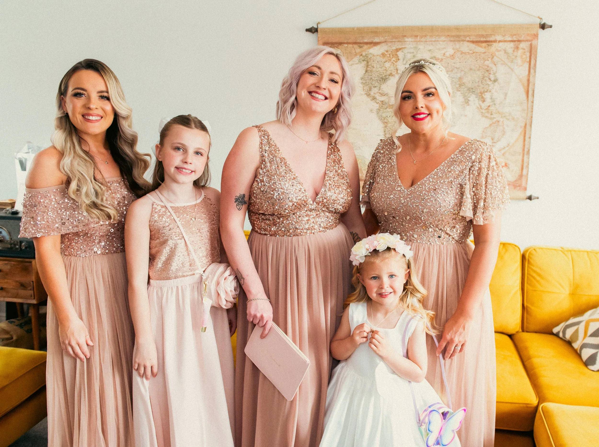 Group of six women and a young girl dressed in pink or white dresses, smiling indoors with a yellow couch and world map in background.