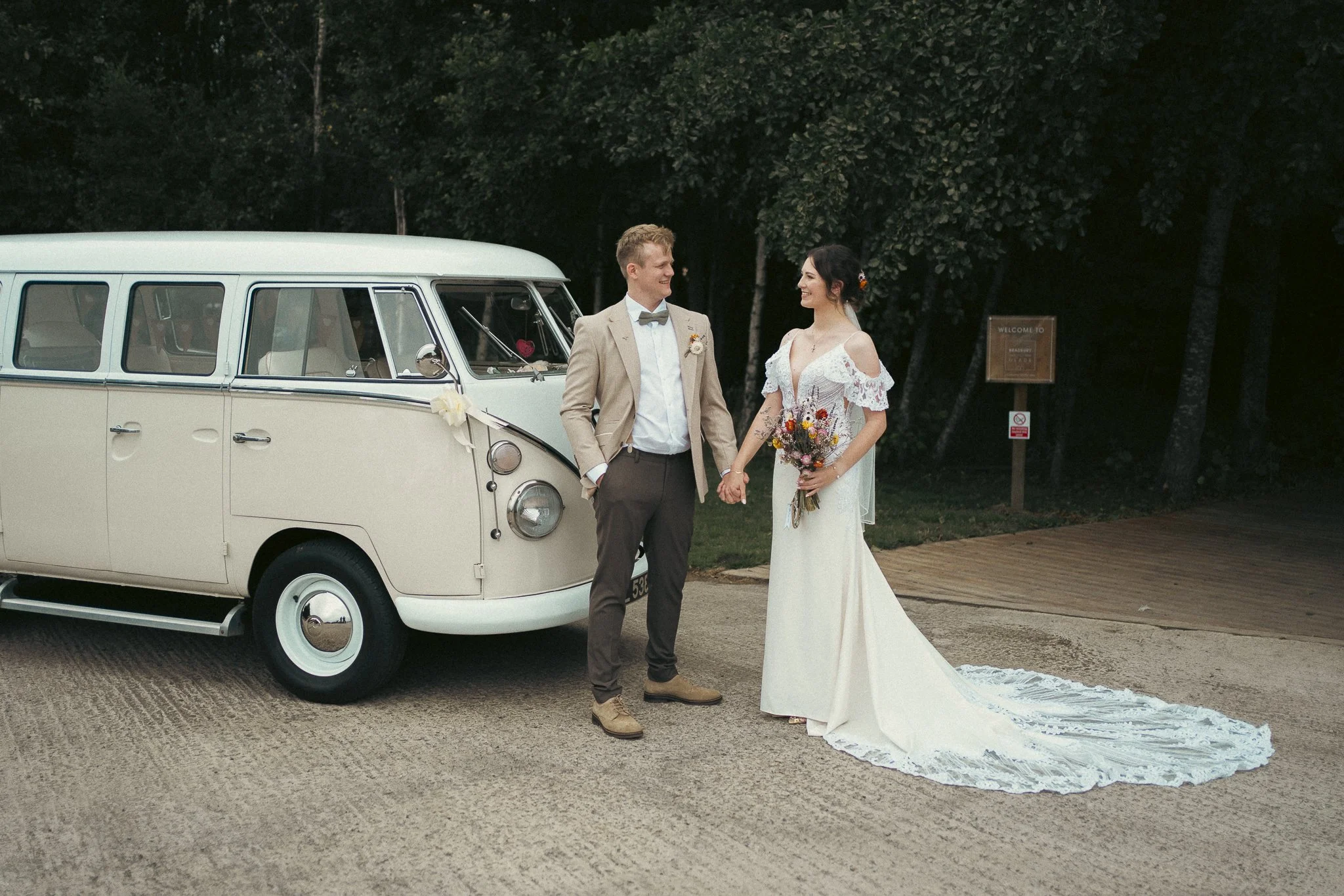 A bride and groom holding hands and smiling at each other outdoors near a vintage cream-colored bus.