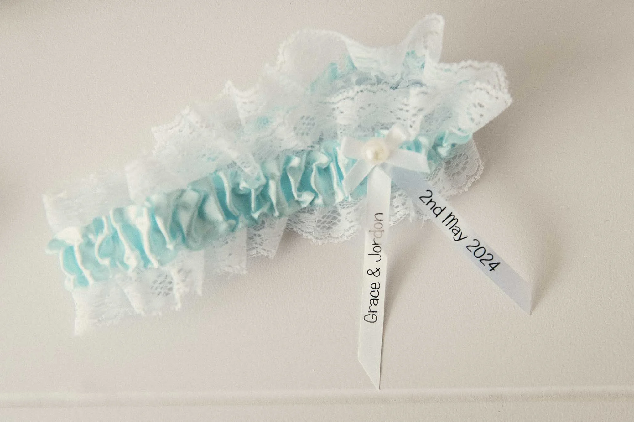 Blue satin and lace wedding garter with a white bow and pearl, with attached ribbons inscribed with names and date.