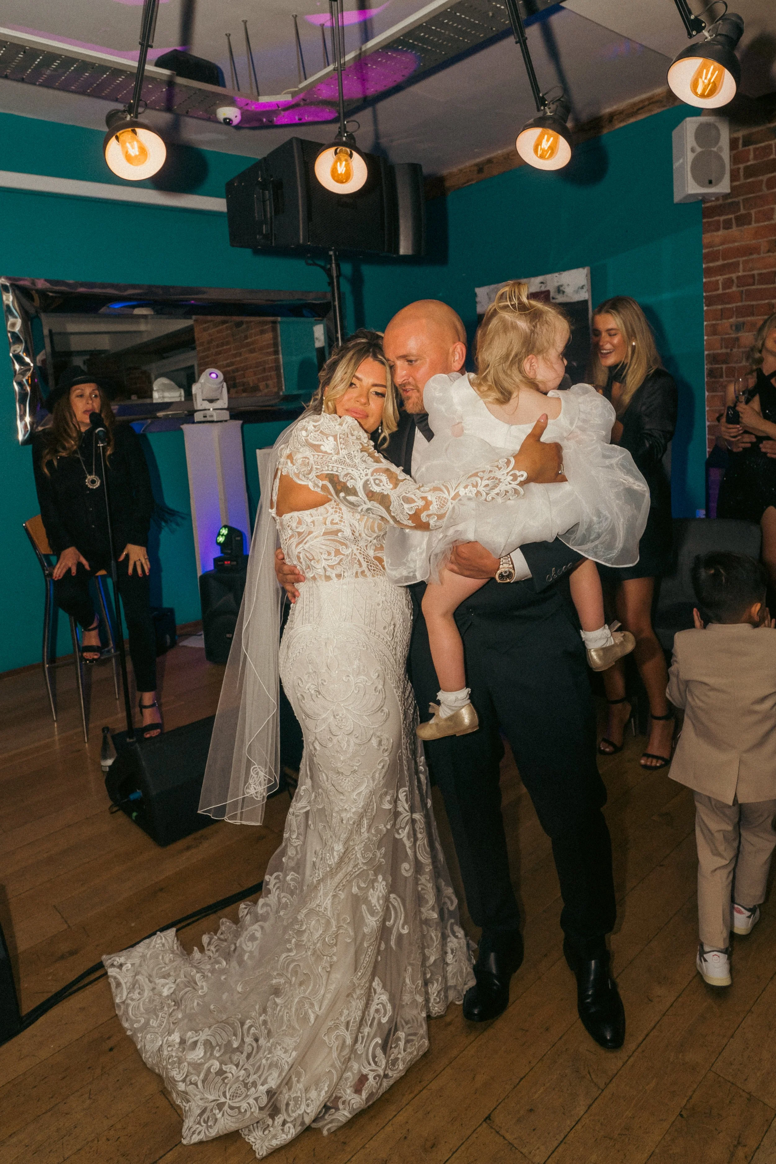 A bride in a white lace wedding dress, a groom in a dark suit, and a young girl in a white dress share a dance at a wedding reception, with guests and a singer performing in the background.