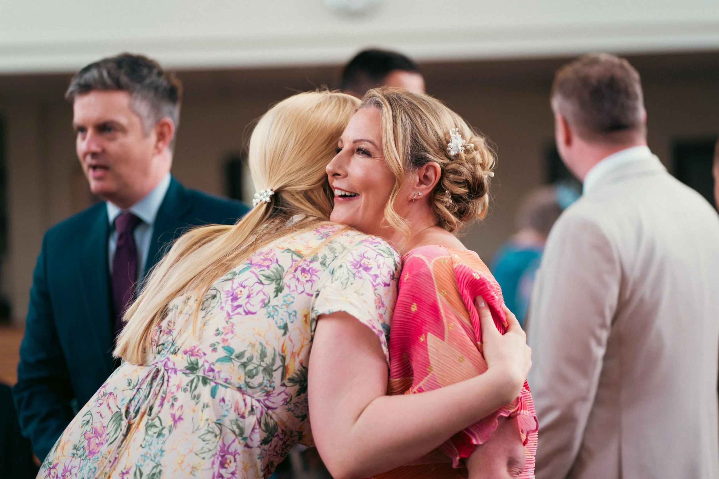 Two women hugging warmly at a social gathering, with smiling faces and people in the background.