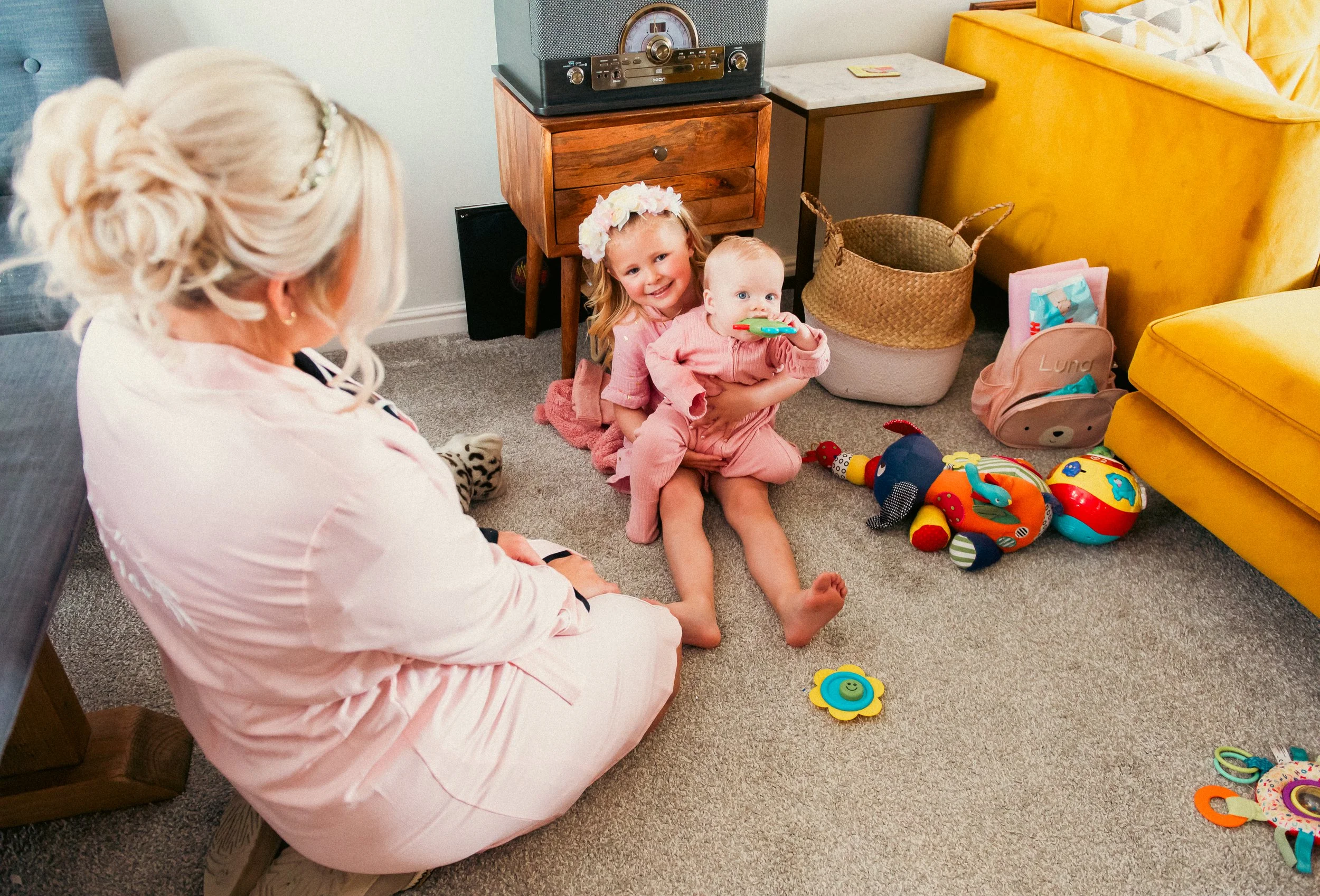 A woman with pink hair and a light pink nurse uniform sitting on the floor, watching two children playing. The children, a young girl wearing a pink outfit and a flower headband, and a baby in pink, are sitting on the carpeted floor with toys around 
