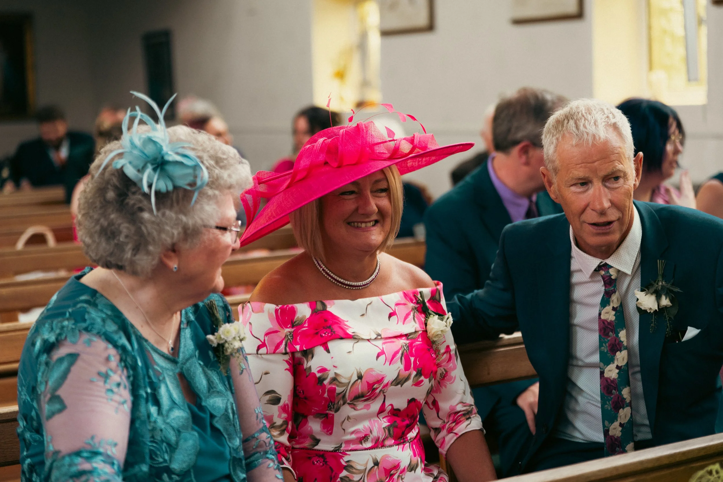 Three people sitting in a church, dressed for a wedding. A woman in the middle is wearing a large pink hat and a pink and white floral dress, smiling. An older woman on the left has curly gray hair and a light blue dress with lace details, and an old