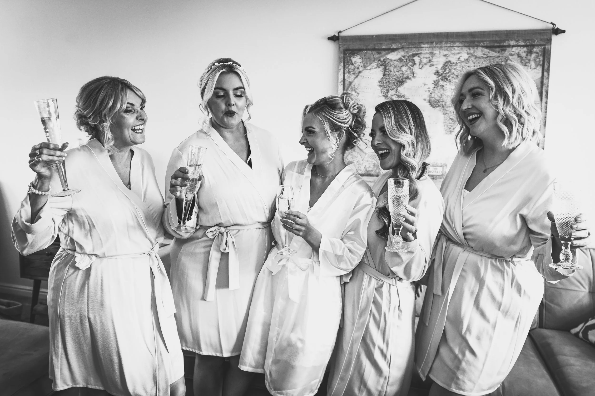 Six women in robes celebrating with champagne in a living room, some smiling and others making playful faces, with a world map on the wall behind them.