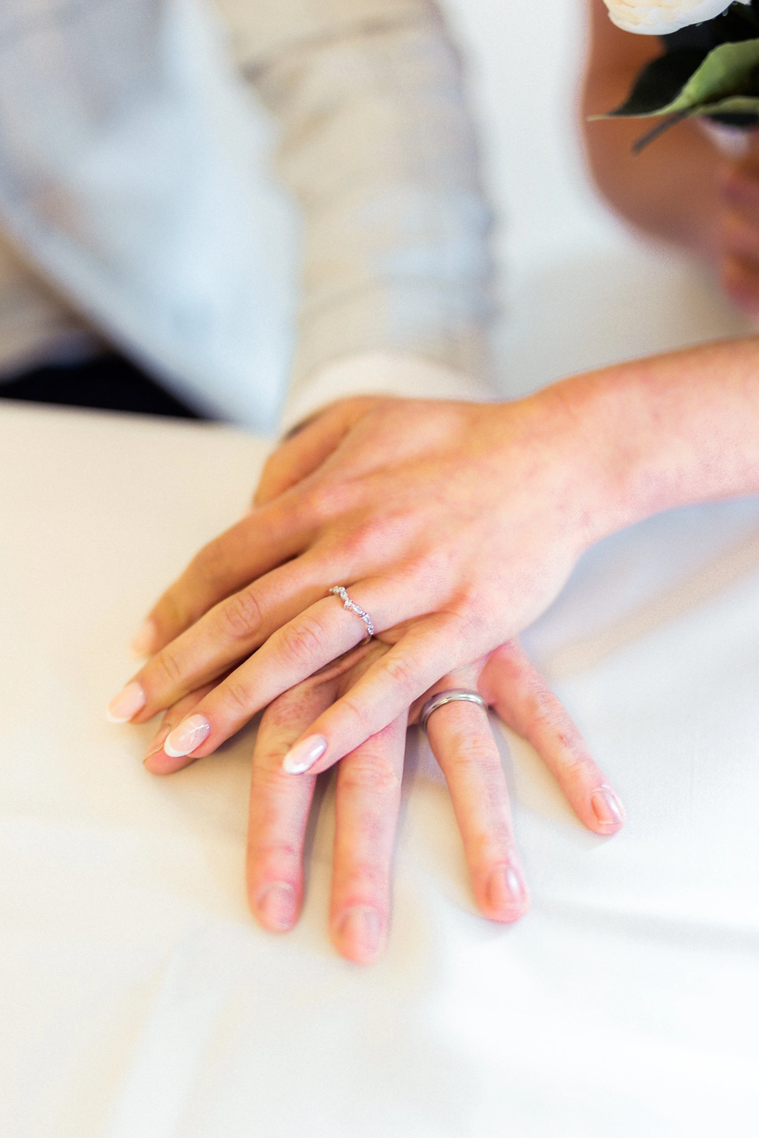 Close-up of a newlywed couple's hands with wedding rings resting on a white surface, with a blurred background of a person's arm and a flower arrangement.