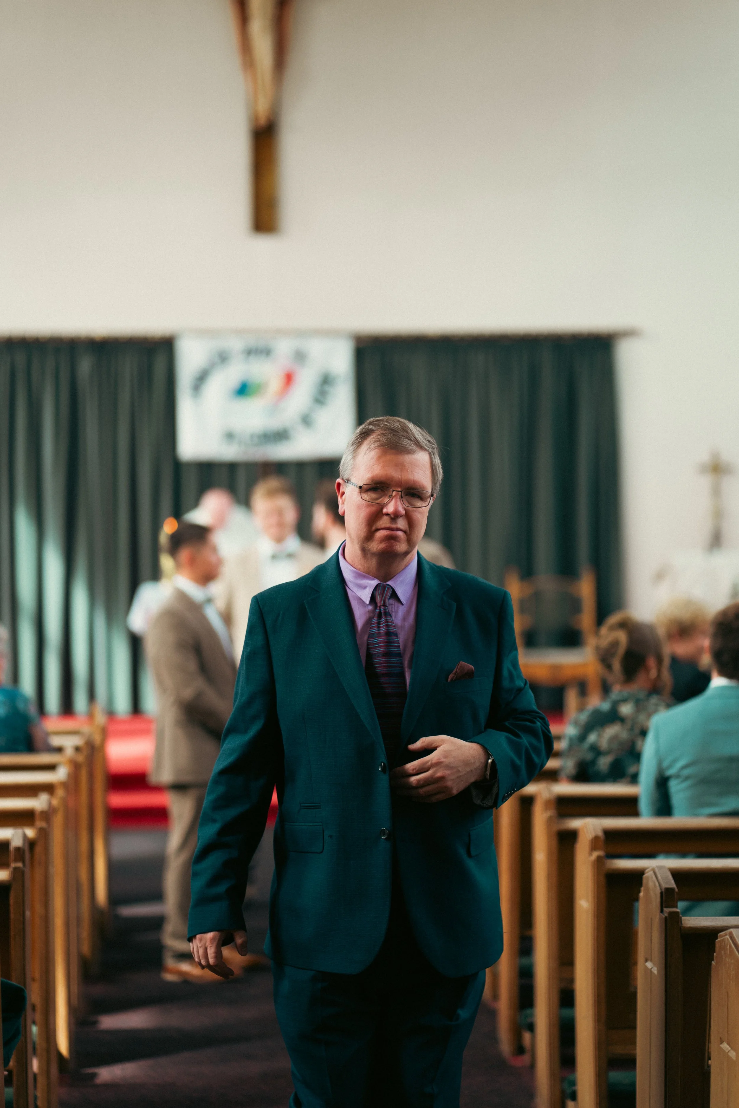 A man in a suit walking down the aisle of a church or ceremony hall with people seated on both sides, and a religious cross on the wall.