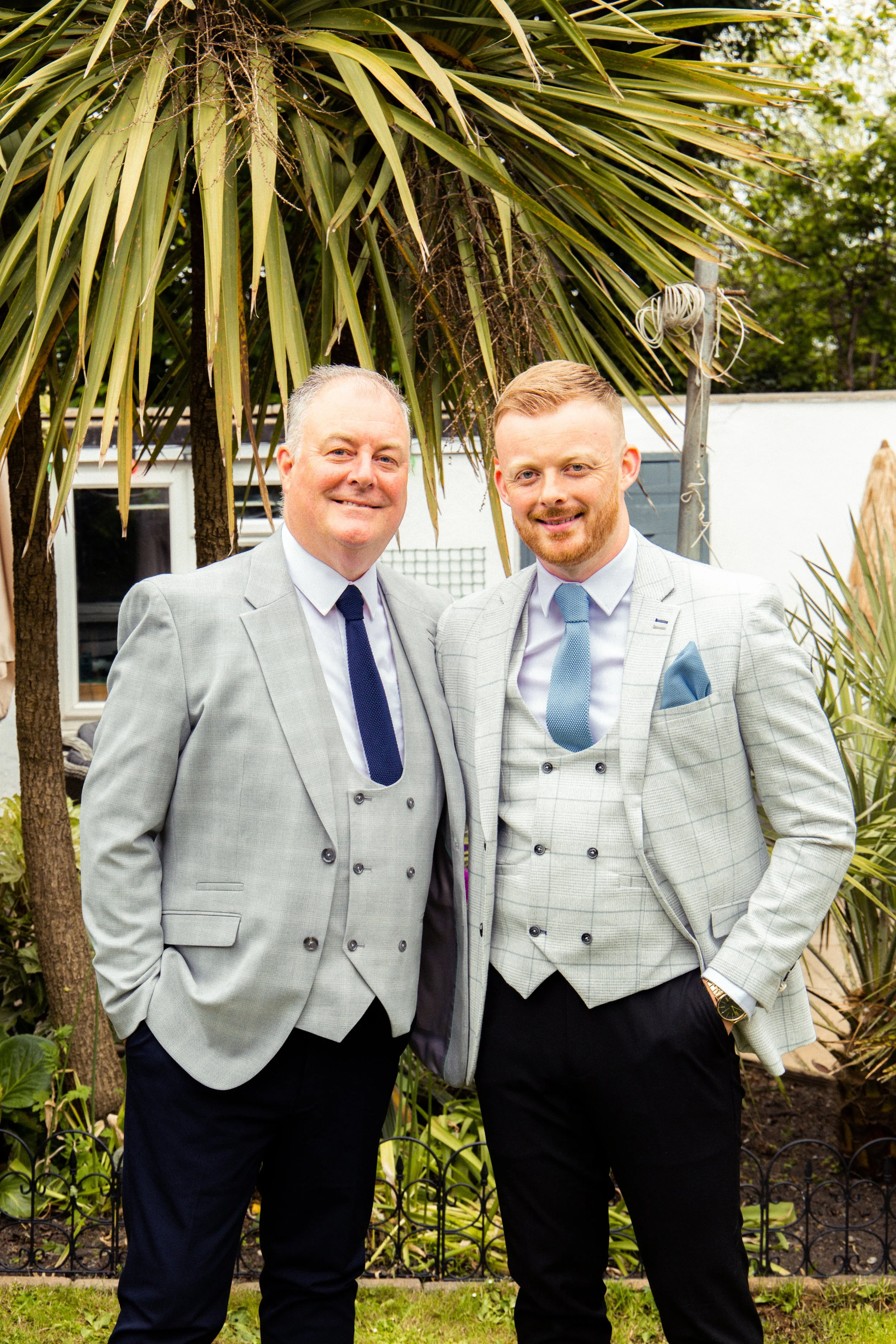 Two men in suits standing outdoors in front of tropical plants, smiling at the camera.