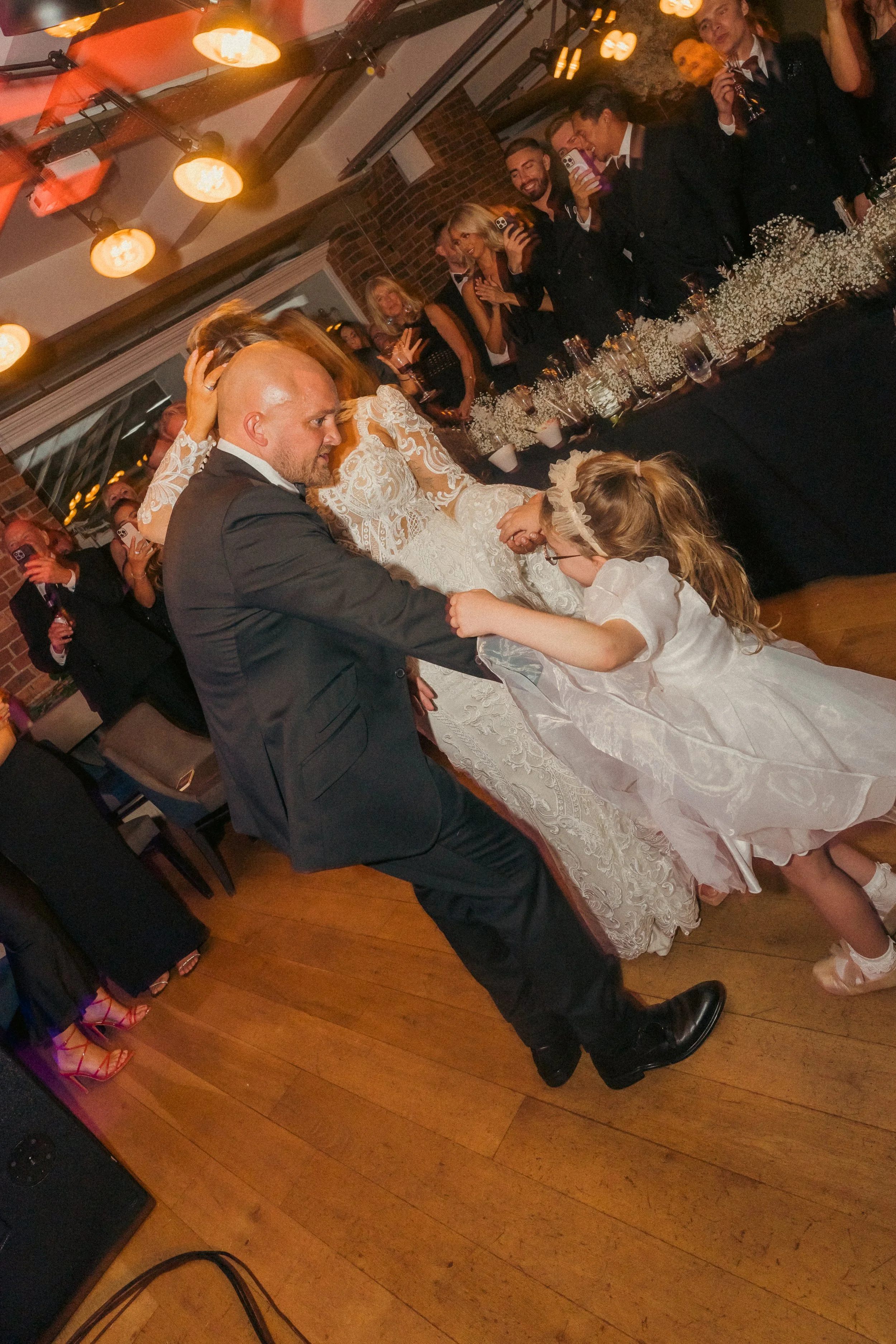 A wedding reception with bride and groom dancing, a girl in a white dress pulling the groom's arm, and guests watching and taking photos.