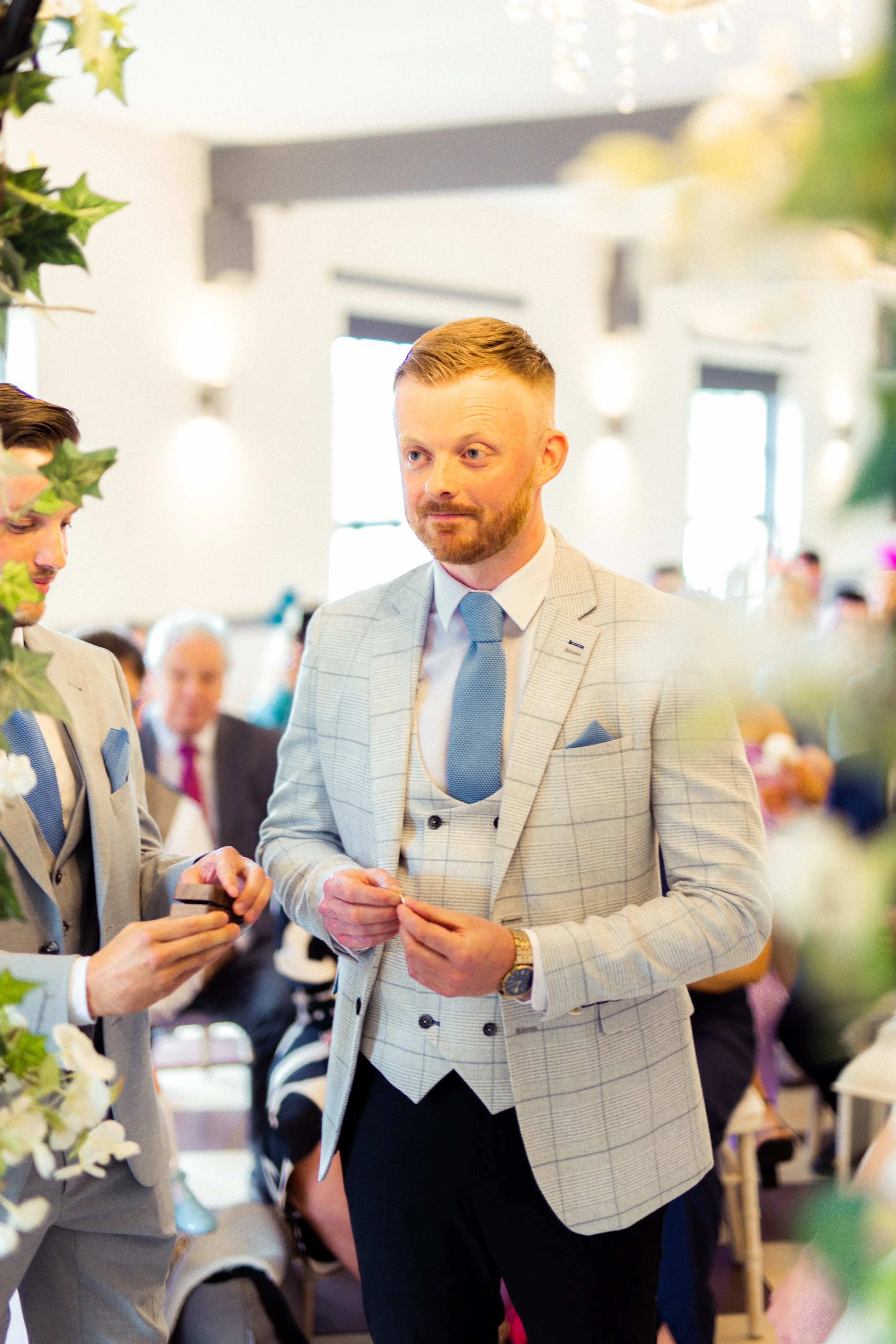 A man with a beard in a light gray checkered suit and blue tie standing at a formal event, possibly a wedding, with other people in suits in the background.