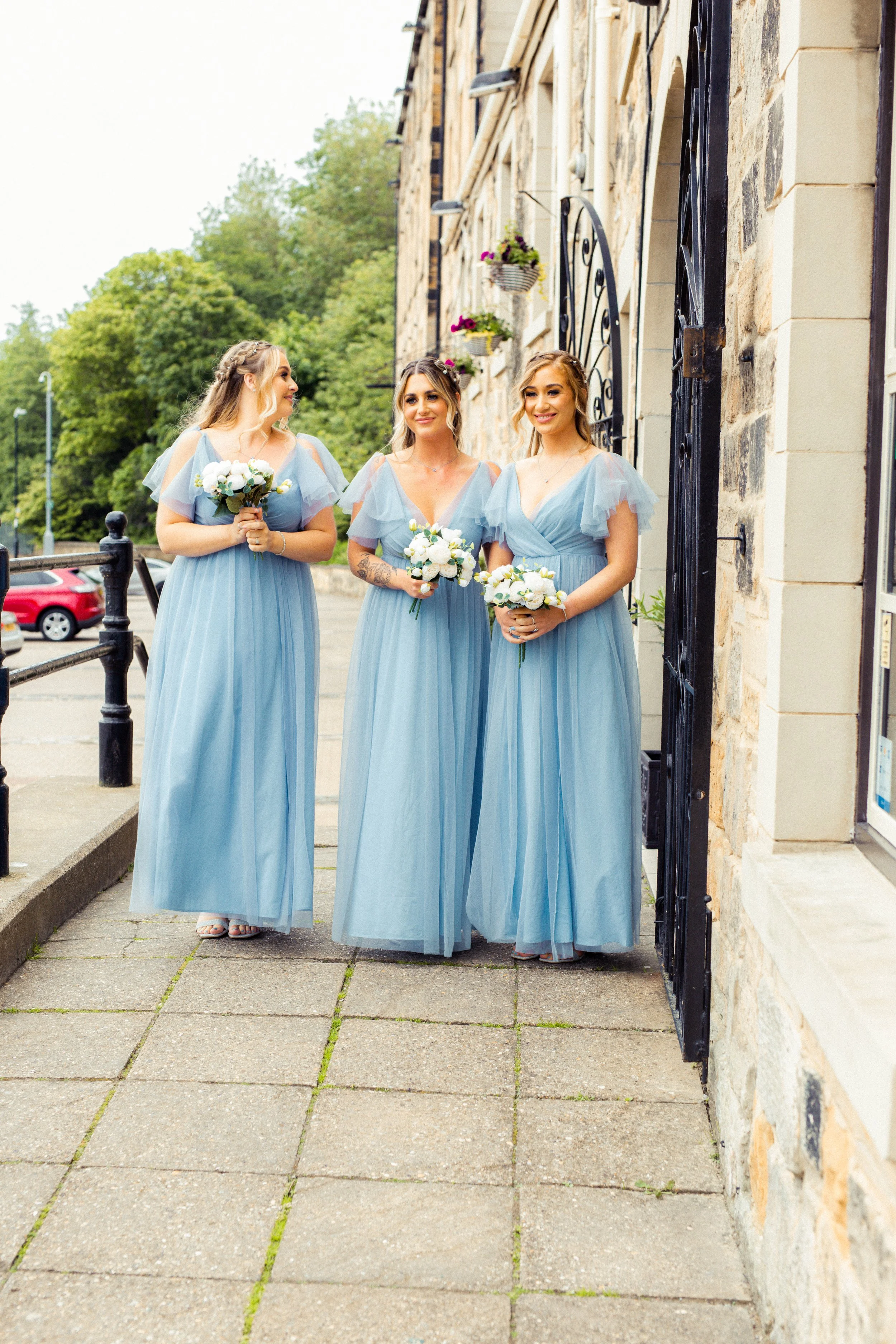 Three women in matching light blue bridesmaid dresses holding bouquets, standing outdoors beside a stone building with a black iron gate.