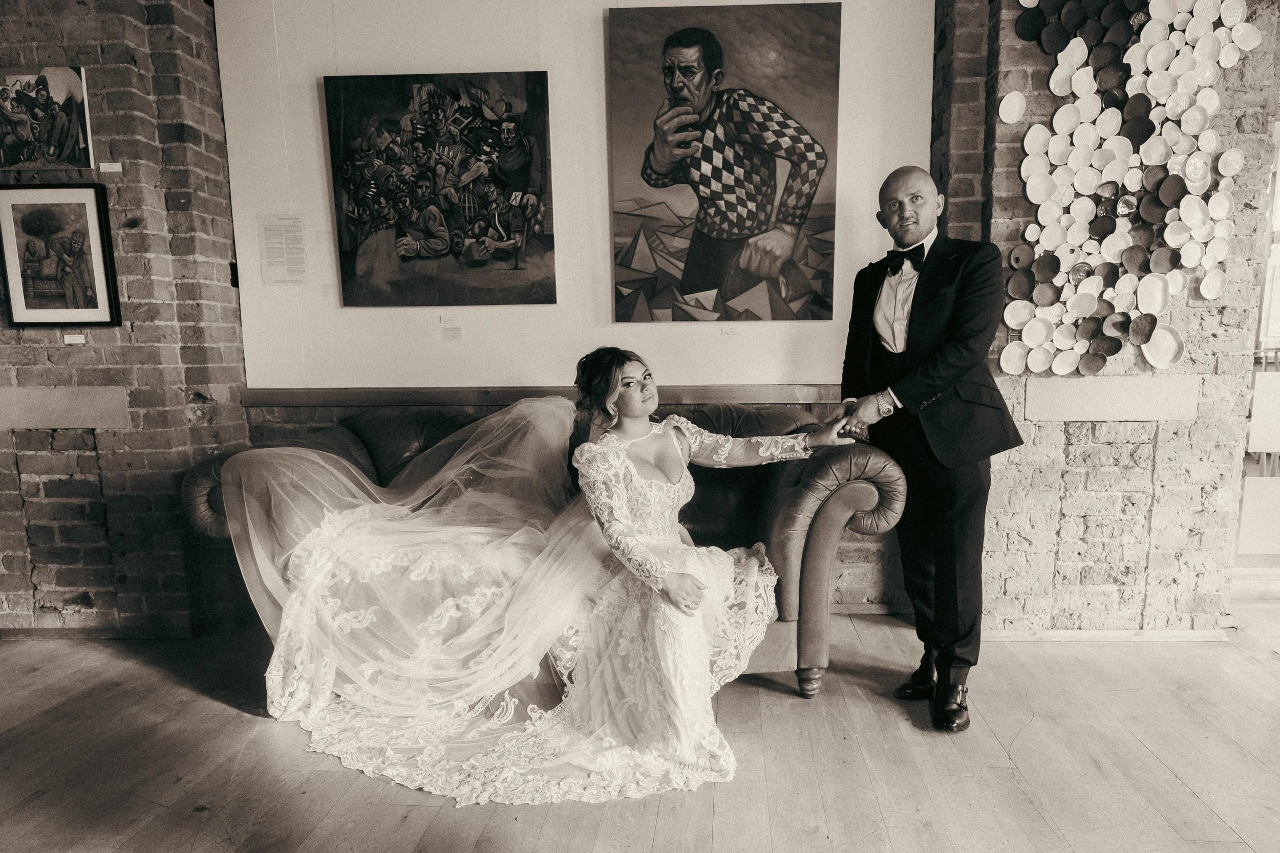 A bride in a lace wedding dress and veil sitting on a vintage brown leather sofa, holding hands with a groom in a tuxedo standing next to her, in an art gallery with brick walls and modern art paintings.