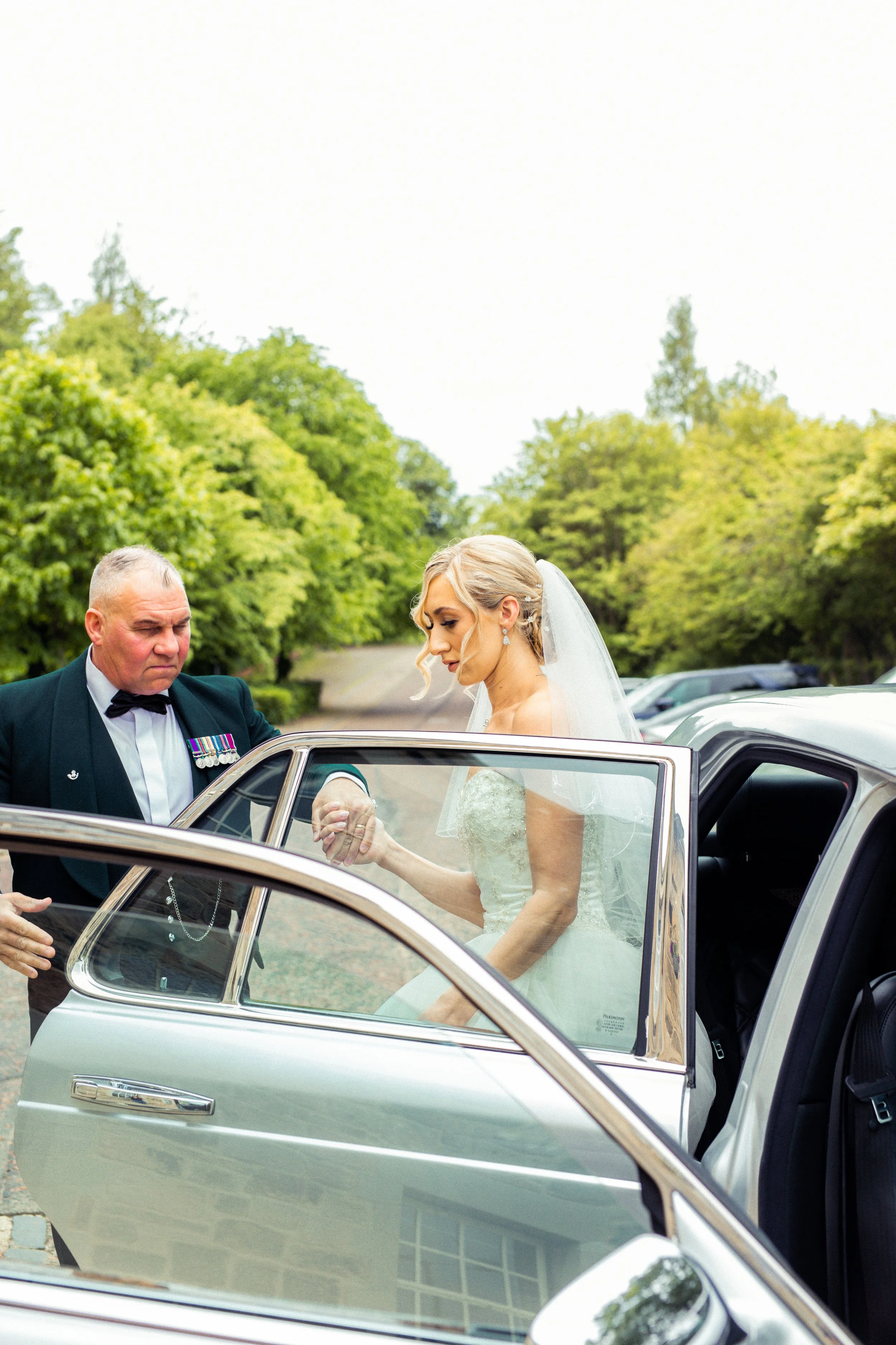 Bride in wedding dress and veil getting into car with man in uniform outdoors.