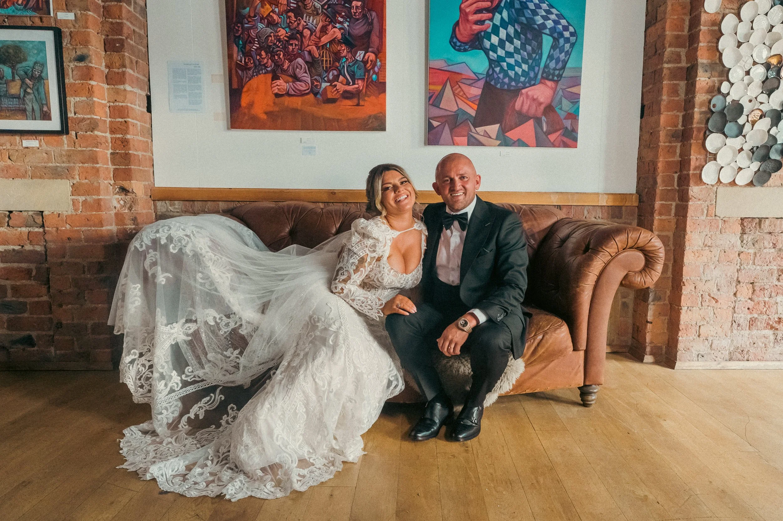 Bride and groom sitting on a leather couch in an art gallery, smiling at the camera. The bride is in a white lace wedding dress with long sleeves, and the groom is in a black tuxedo. Behind them are colorful paintings on brick walls.