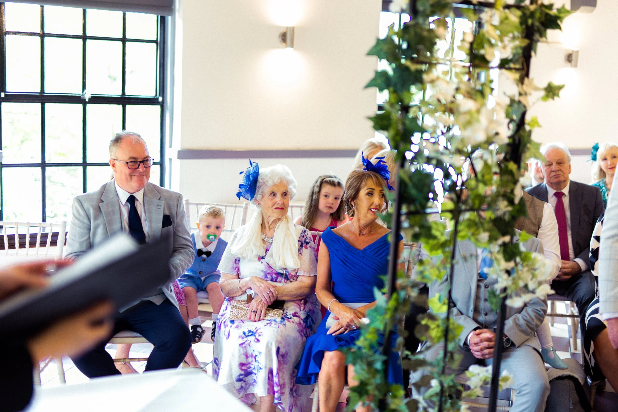 Group of people seated in a well-lit indoor space with large windows, attending a formal event or wedding, decorated with greenery and white flowers.