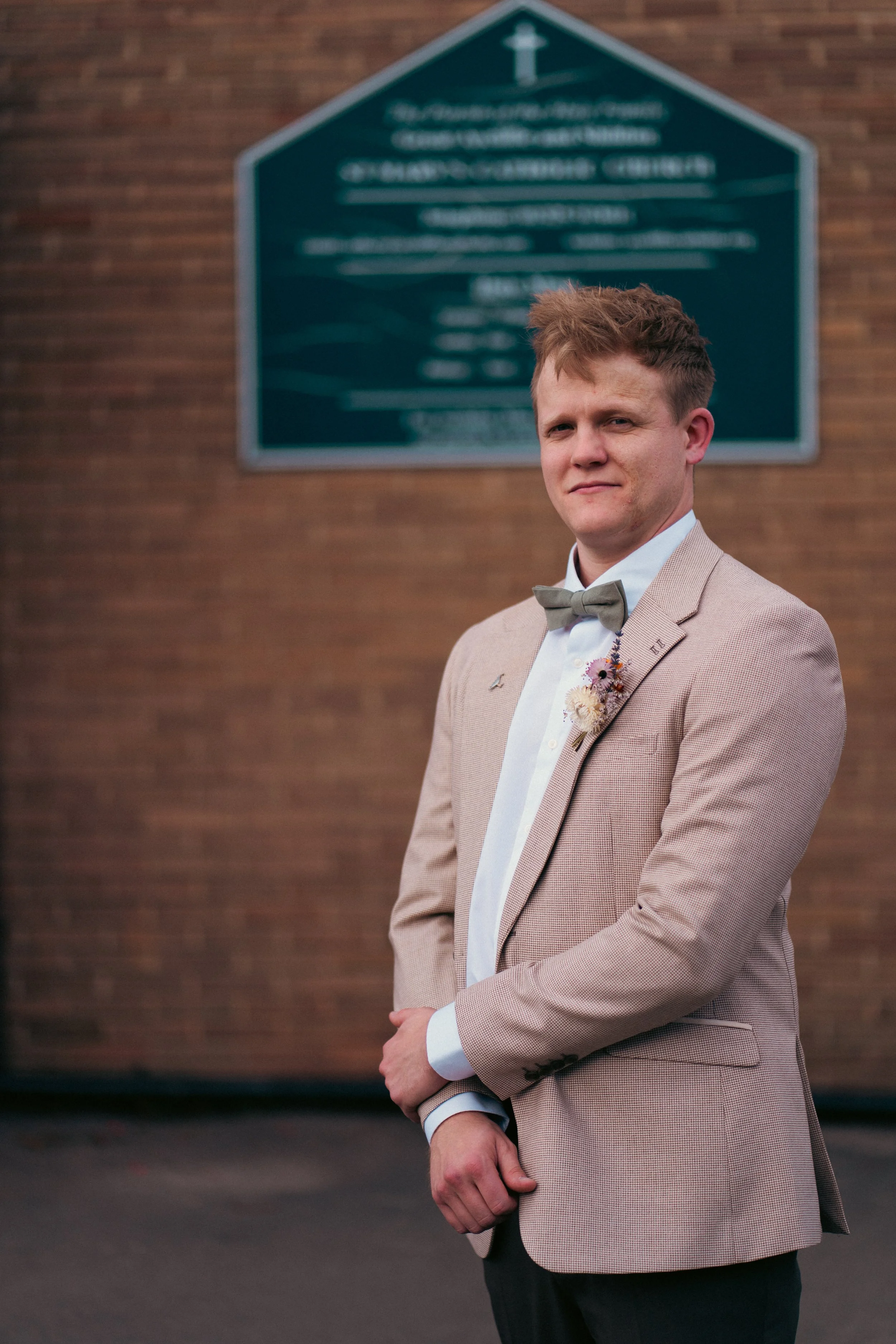 A young man in a beige suit and green bow tie standing outdoors in front of a brick wall with a blurred green church sign in the background.
