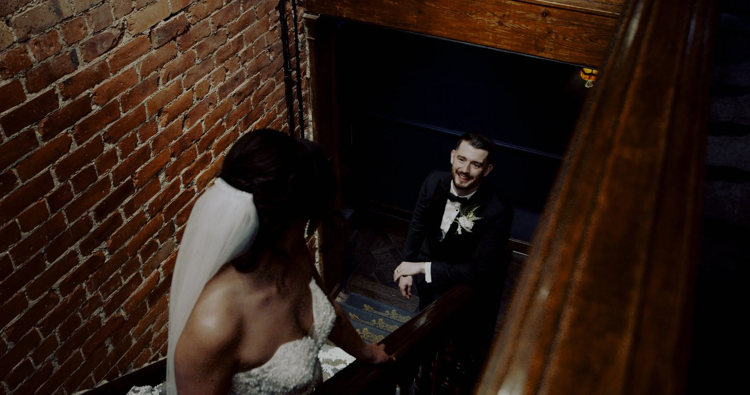 A bride with dark hair and a white gown looking down a staircase at a groom in a black tuxedo and bow tie, who is smiling up at her. The setting has exposed red brick walls and dark wood accents.