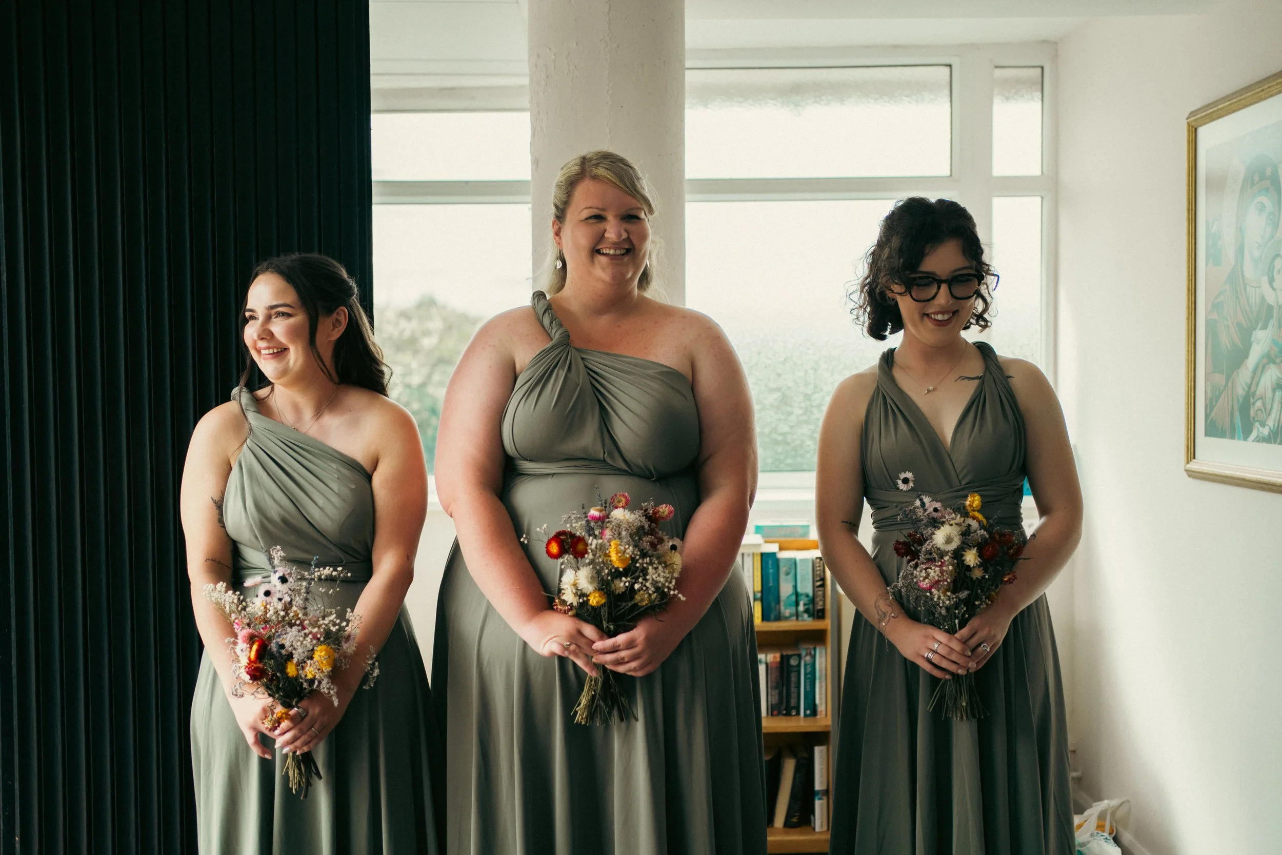 Three women in matching grey bridesmaid dresses holding bouquets of wildflowers, standing indoors near a window with bookshelves and artwork in the background.