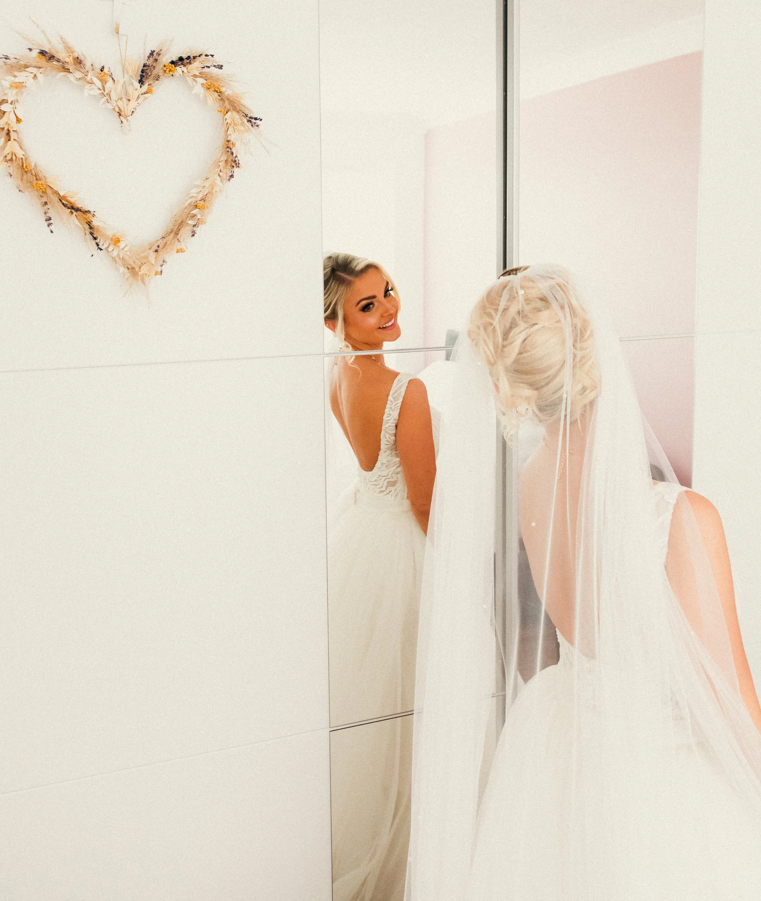 A bride looking into a mirror with her reflection showing her smiling face, her wedding dress and veil, with a decorative heart-shaped floral arrangement on the wall.