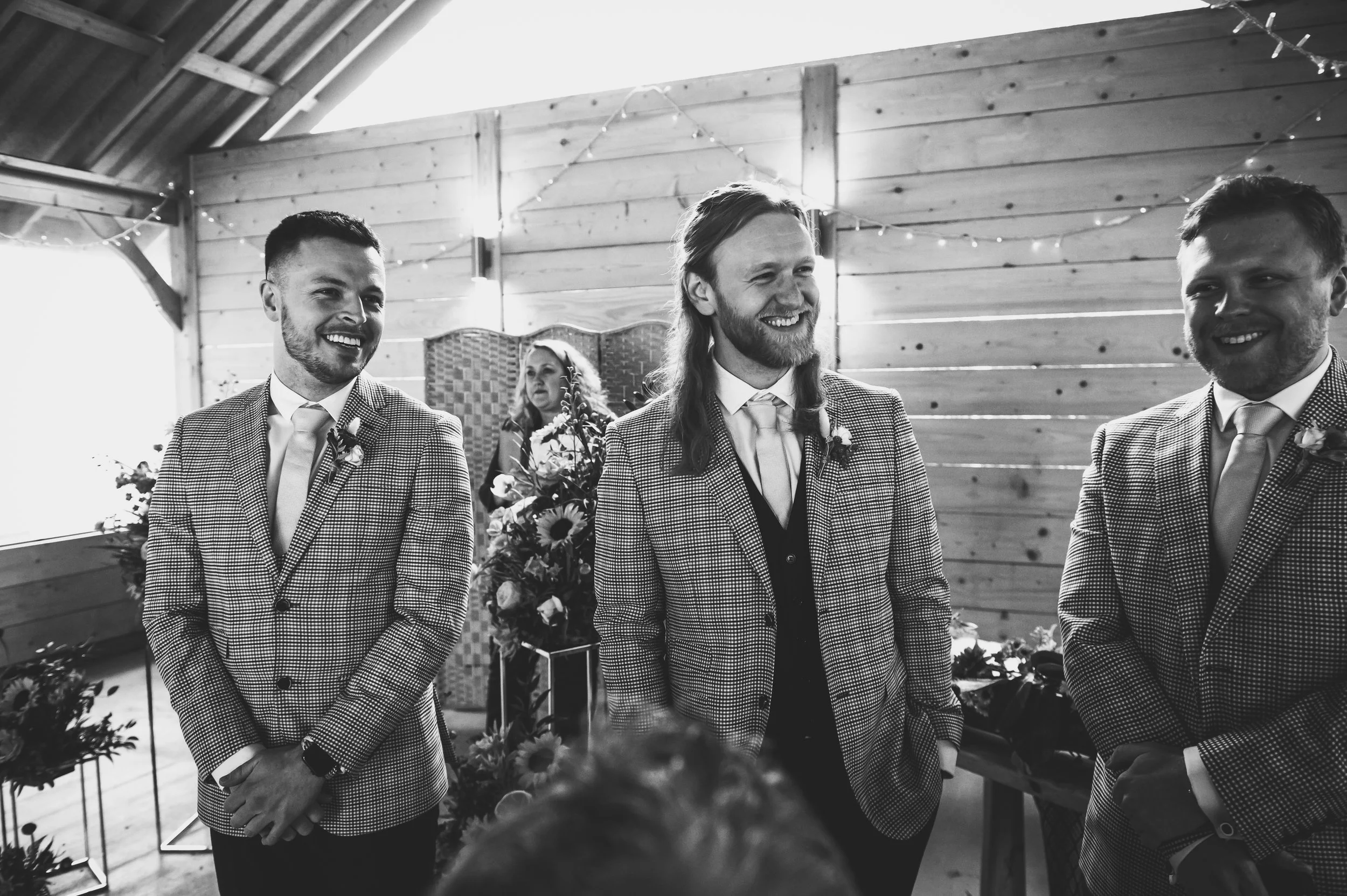 Black and white photo of three men wearing checked suits and ties at a wedding, smiling and standing in a wooden venue with string lights, while a woman with long hair and a floral bouquet stands in the background.