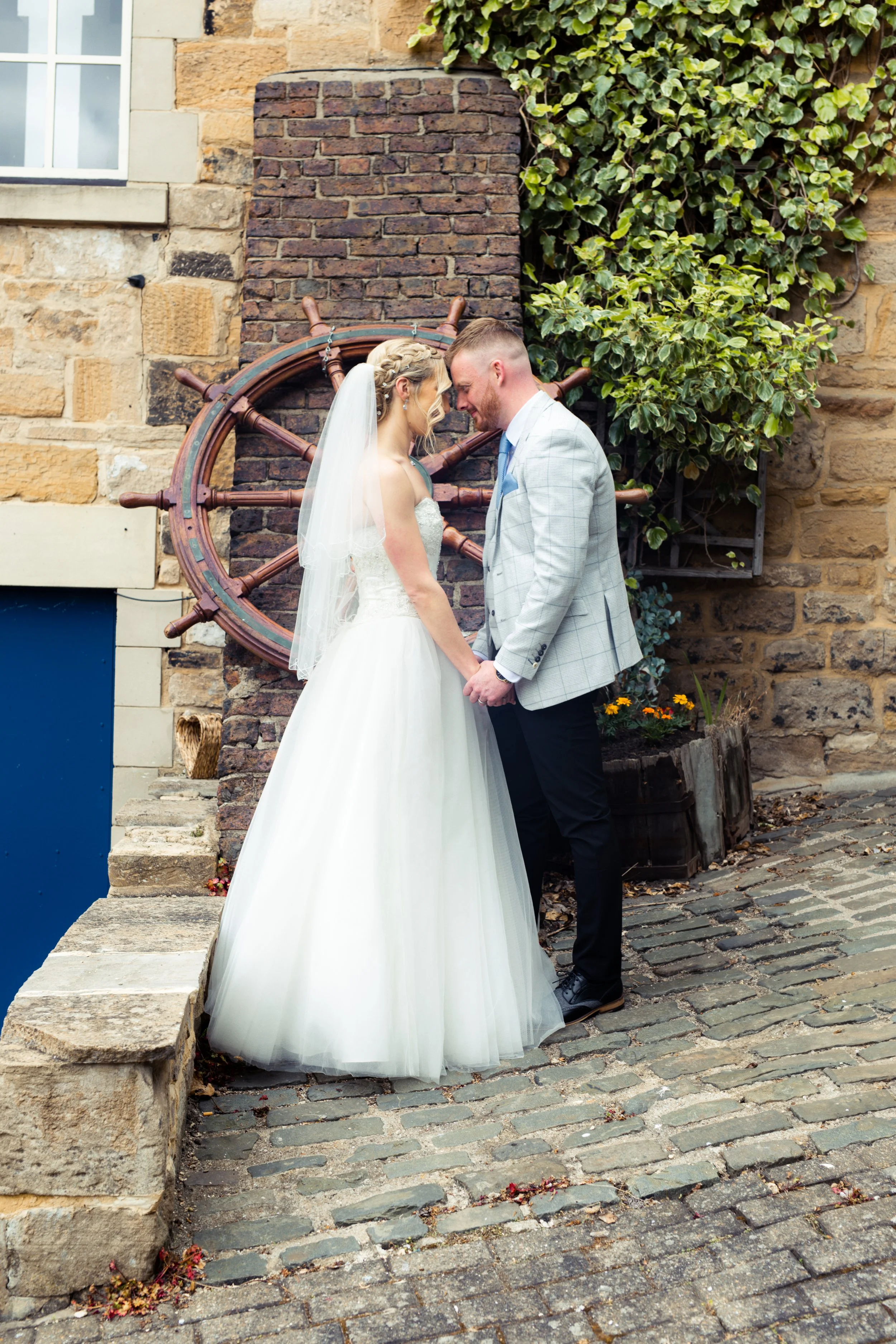 A bride and groom standing close, holding hands and touching foreheads, outdoors on a cobblestone street with a brick wall and a ship's wheel in the background.
