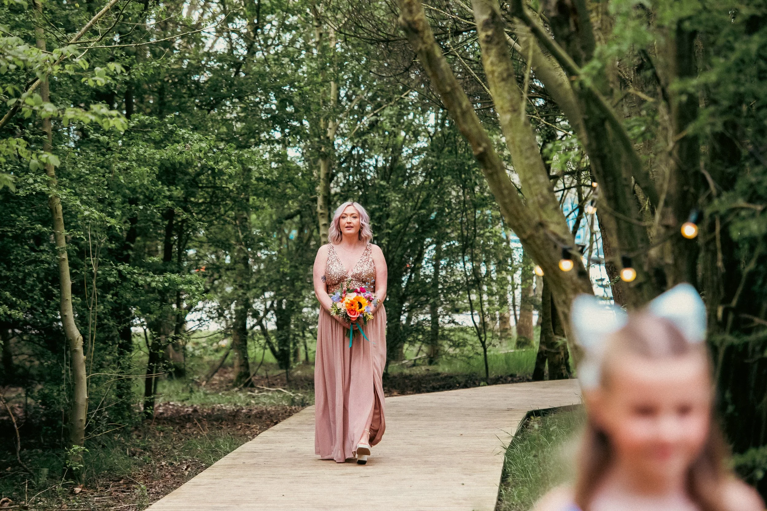 Woman in a long dress holding a colorful flower bouquet walking along a wooden pathway in a wooded area