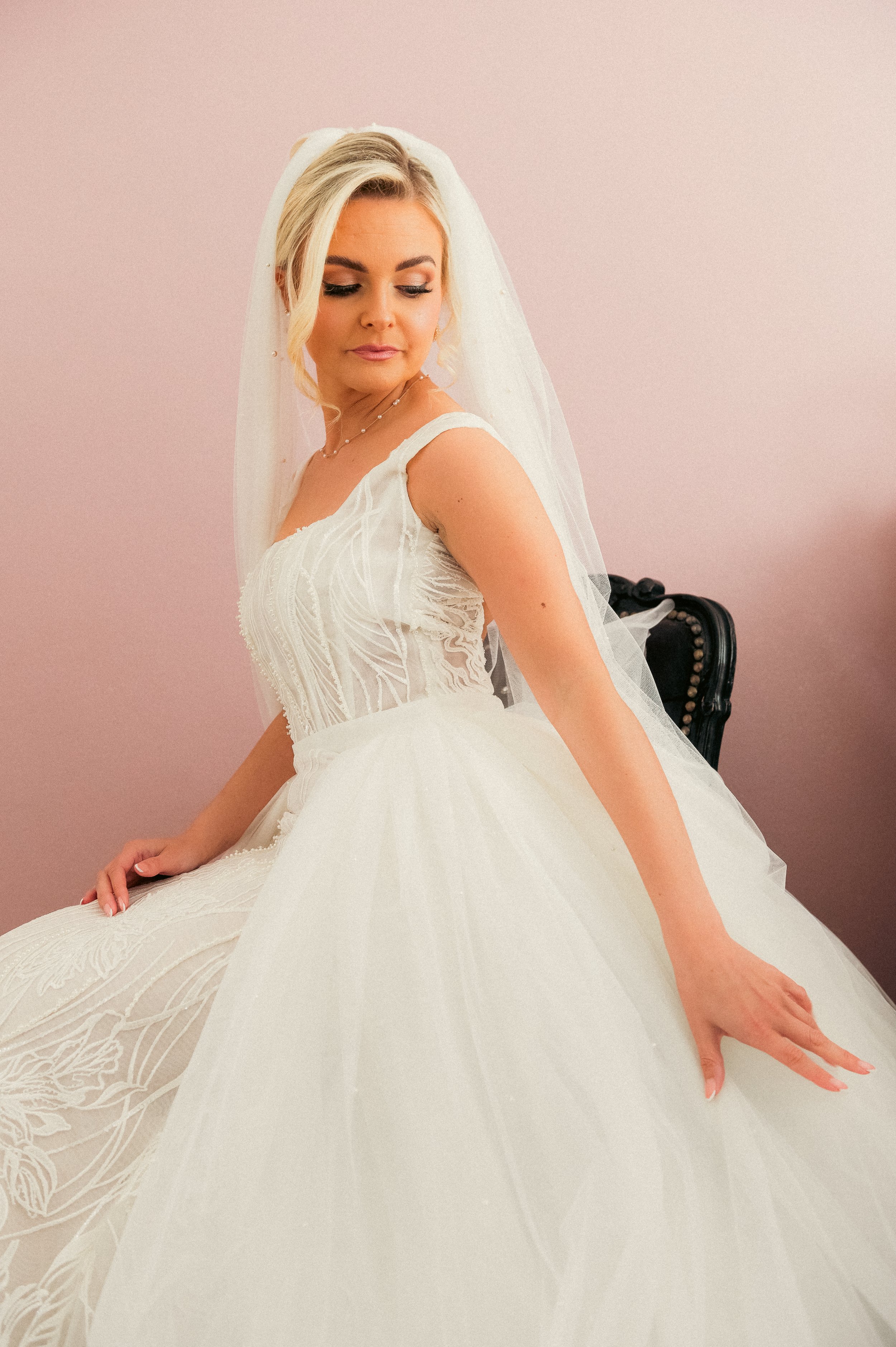 A bride in a white wedding gown with lace details, a veil, and a necklace, sitting on a dark chair with a pink wall in the background.