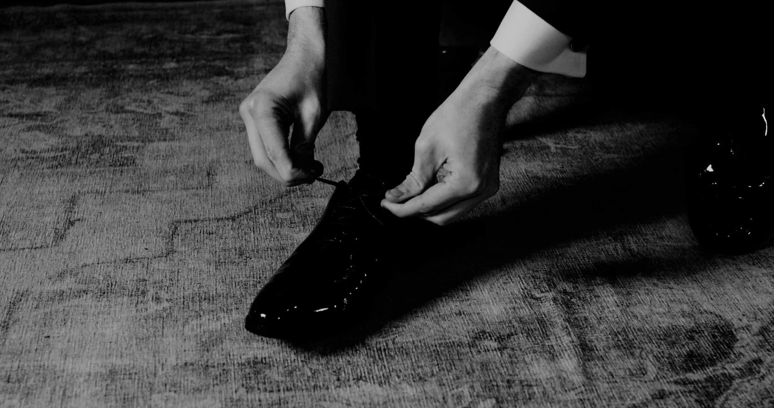 Person in formal attire tying shoelaces of a polished black dress shoe on a wooden floor.