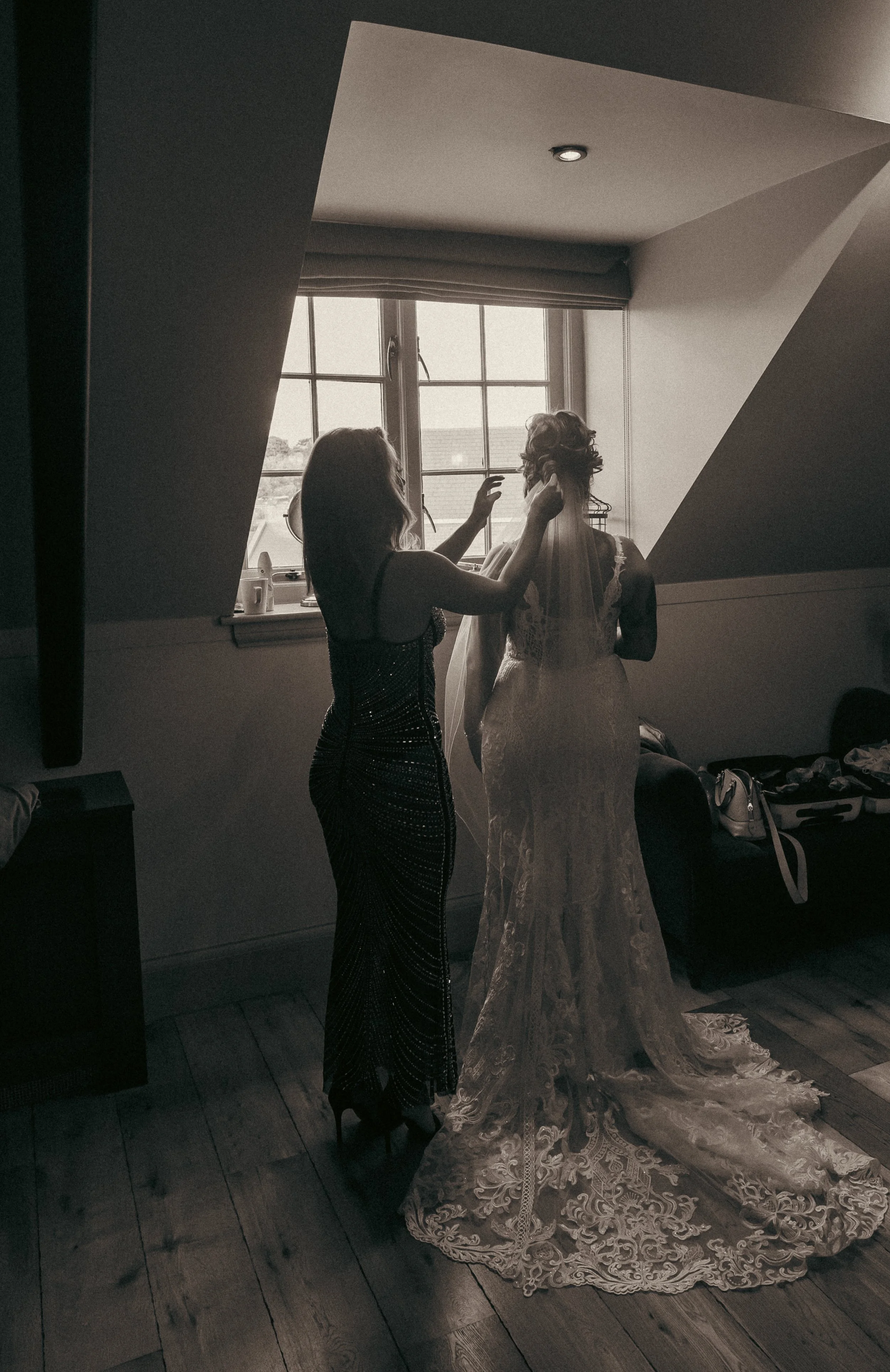 A woman in a lace wedding dress is being prepared by another woman in a black dress in a dimly lit room with a window.