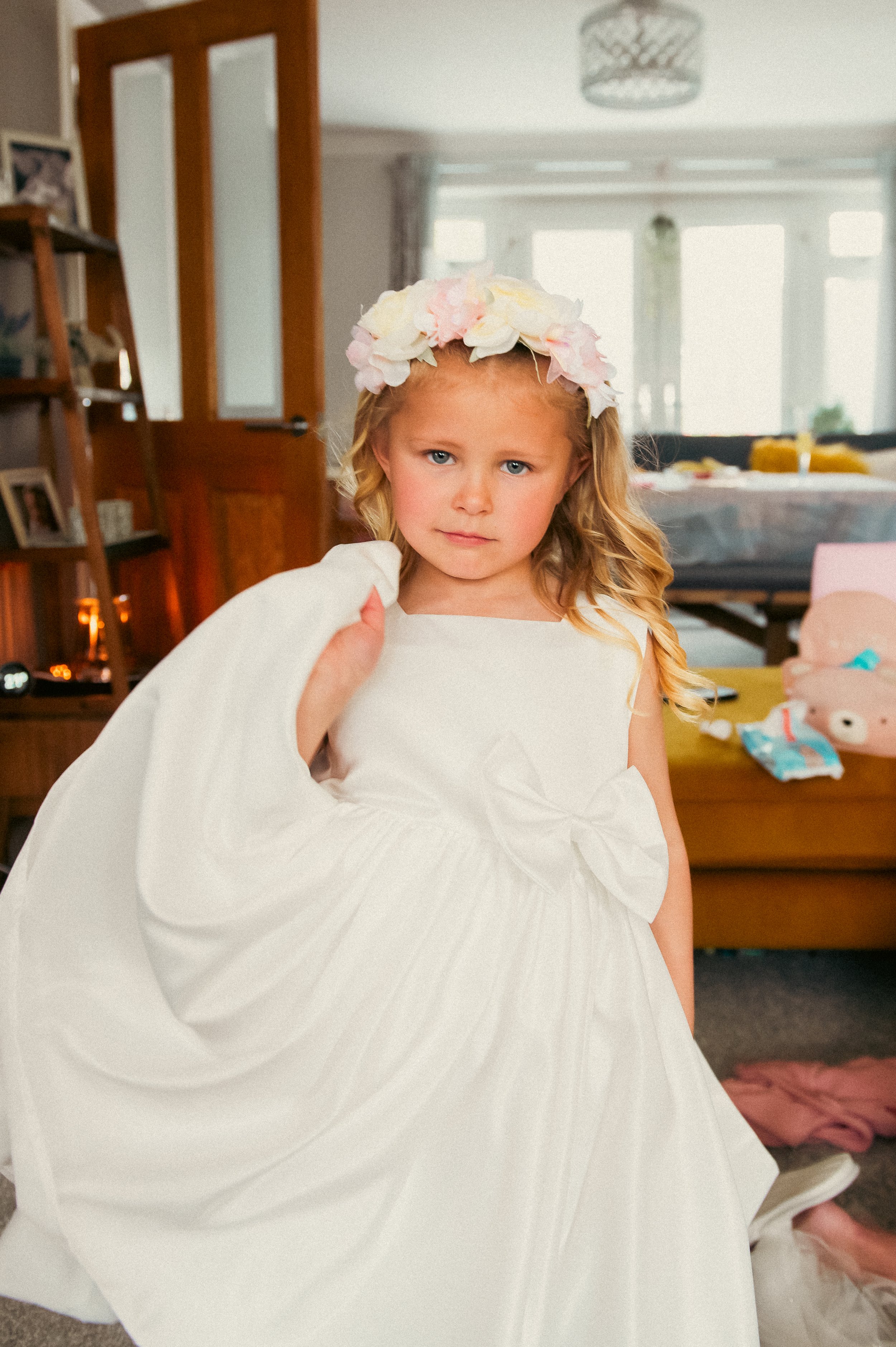 A young girl with blonde hair and blue eyes wearing a white dress with a large bow, and a floral headband of pink and white flowers. She is standing indoors in a room with a wooden bookshelf, a brown door, and a yellow piece of furniture in the backg