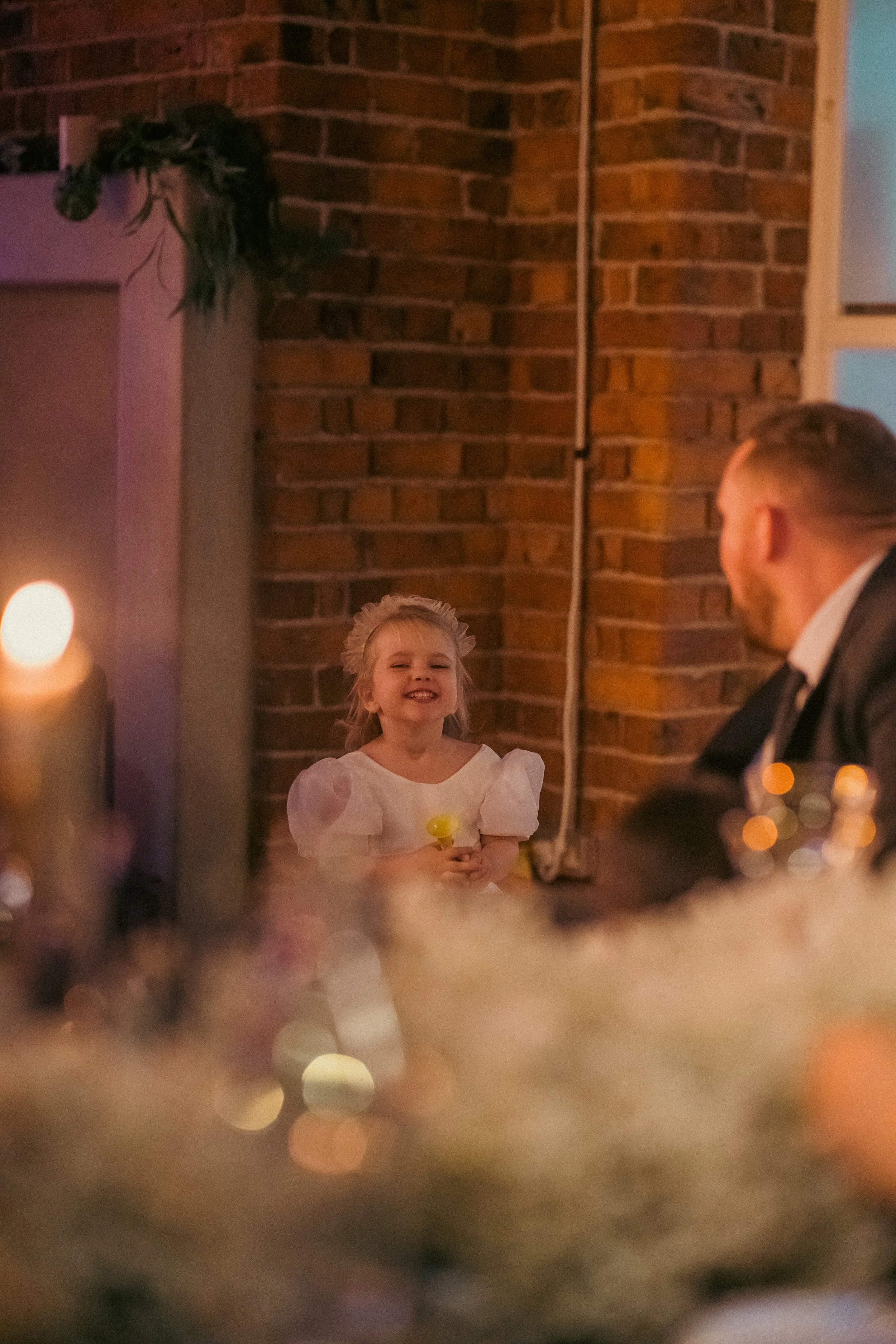 A young girl with a big smile, wearing a white dress with puffed sleeves and a headband, stands in front of a brick wall holding a yellow toy or object, while a man dressed in a suit sits nearby in an indoor setting with warm lighting and blurred for