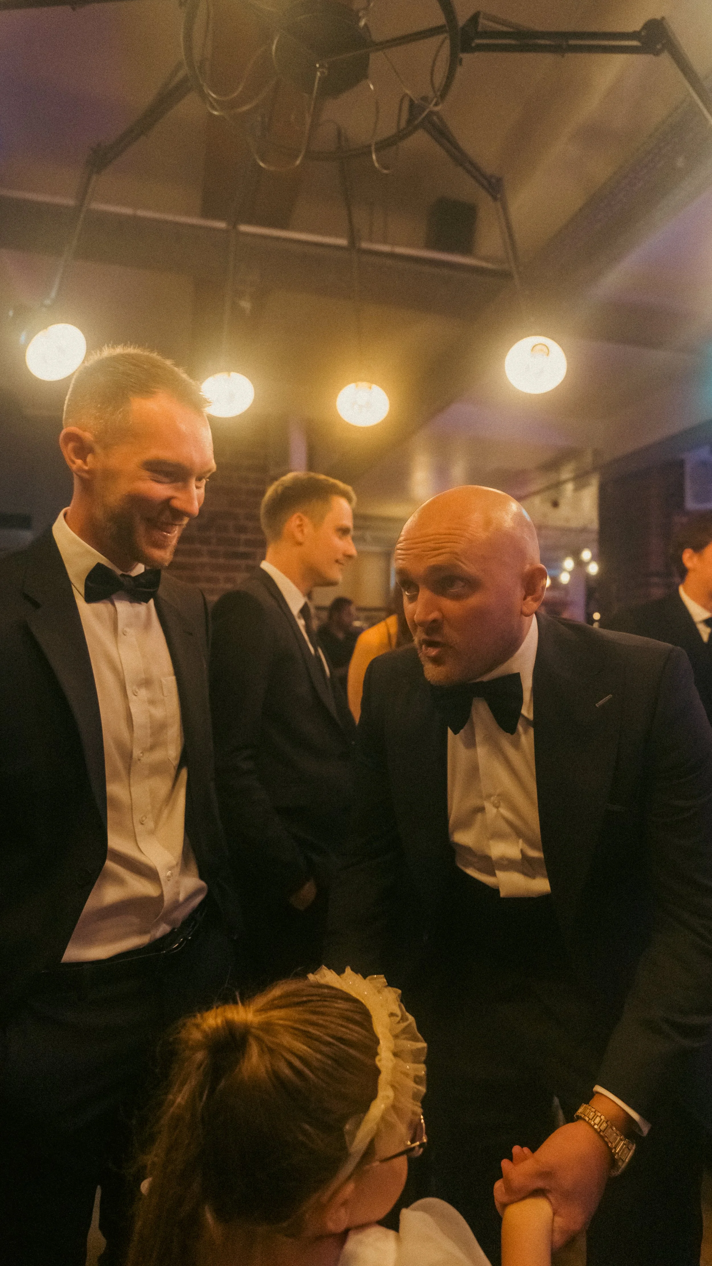 Three men dressed in tuxedos and bow ties talking to a young girl with glasses and a headband at a formal indoor event with warm lighting.