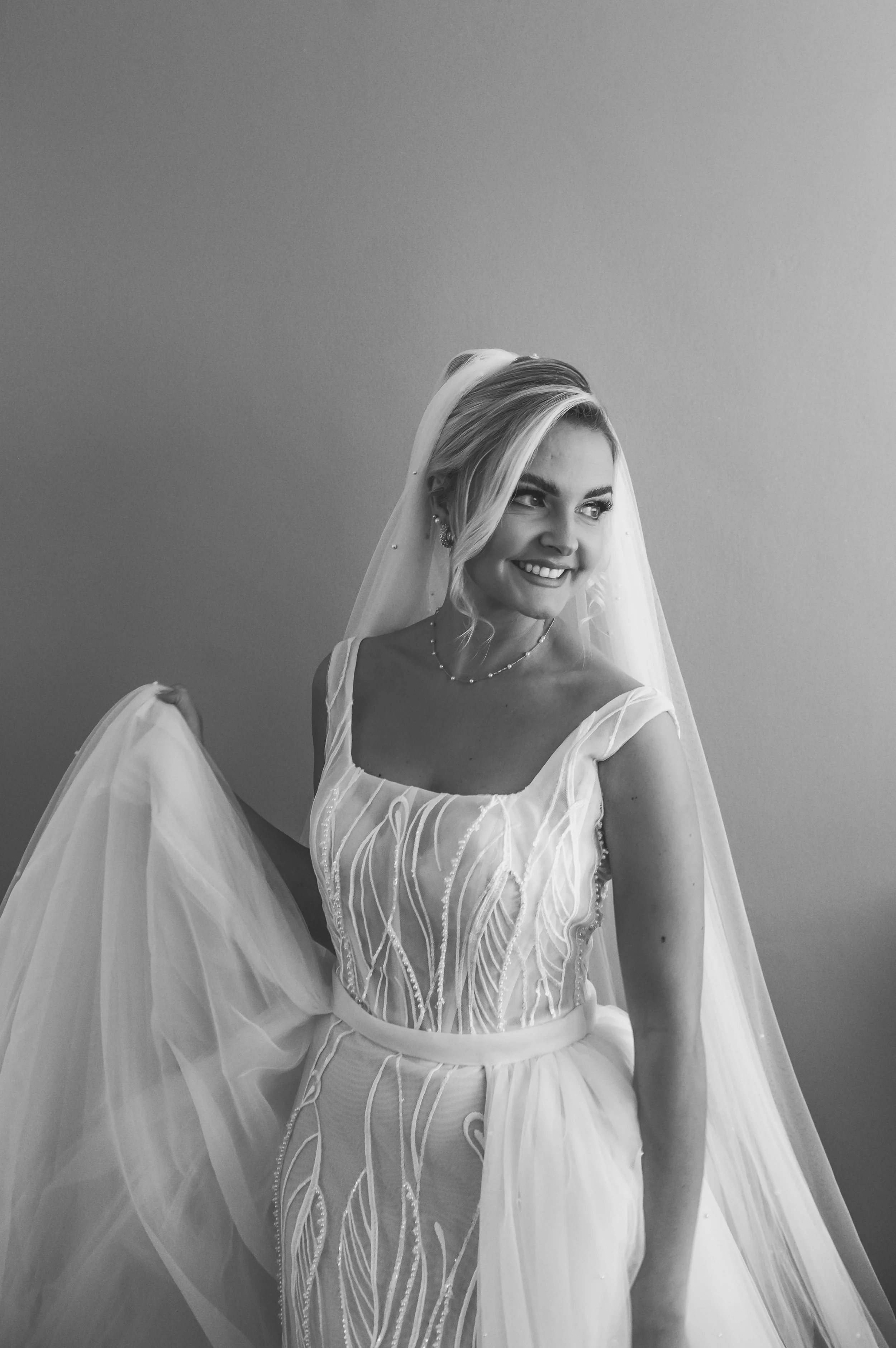 Black and white photo of a smiling woman in a wedding dress with a veil, holding part of her dress, standing against a plain wall.