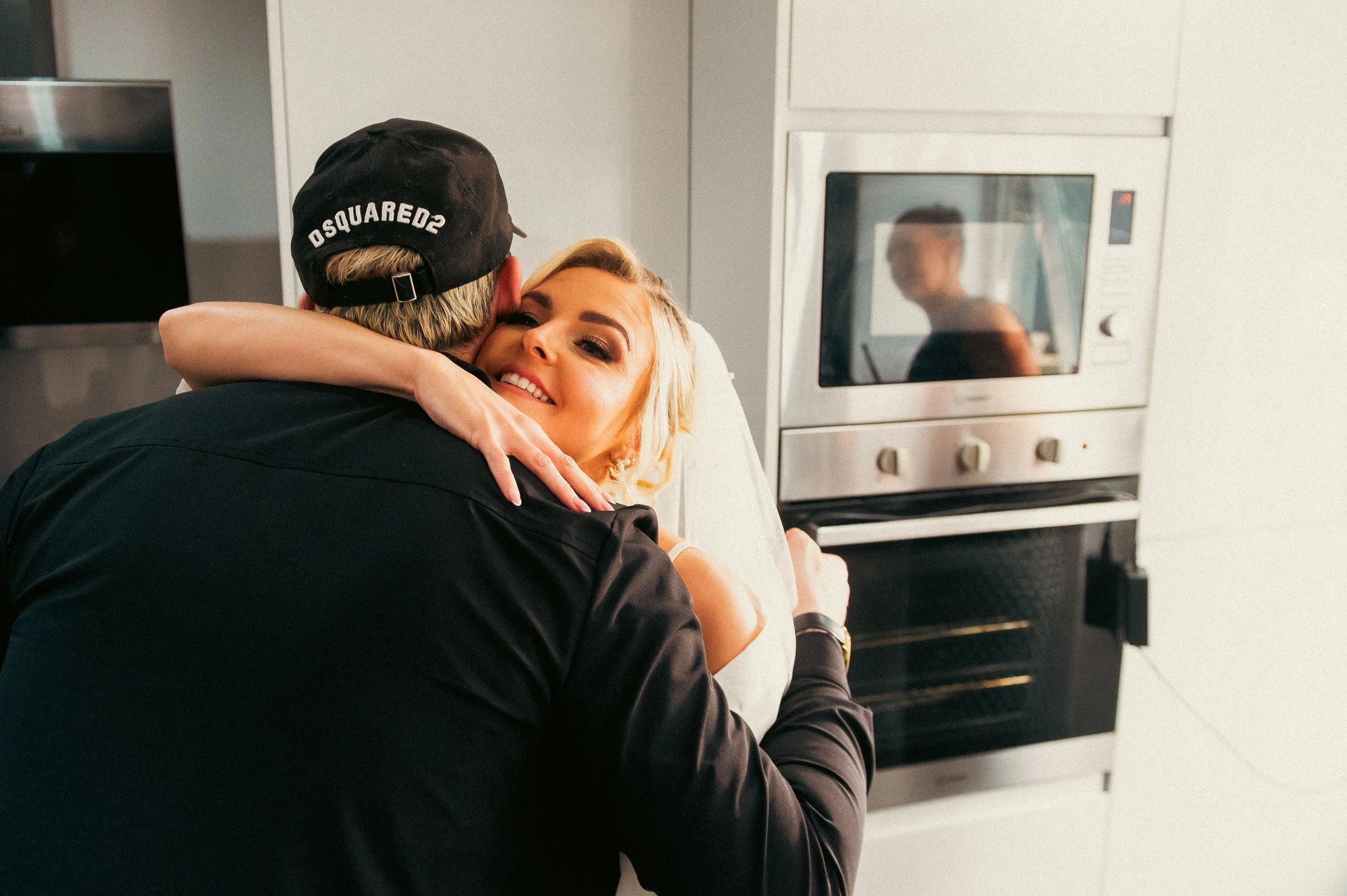 A woman with blonde hair smiling and hugging a man wearing a black cap and jacket in a kitchen. The woman is lying in a bed with a white headboard. In the background, there is a stainless steel oven with a reflective glass door and a person visible t