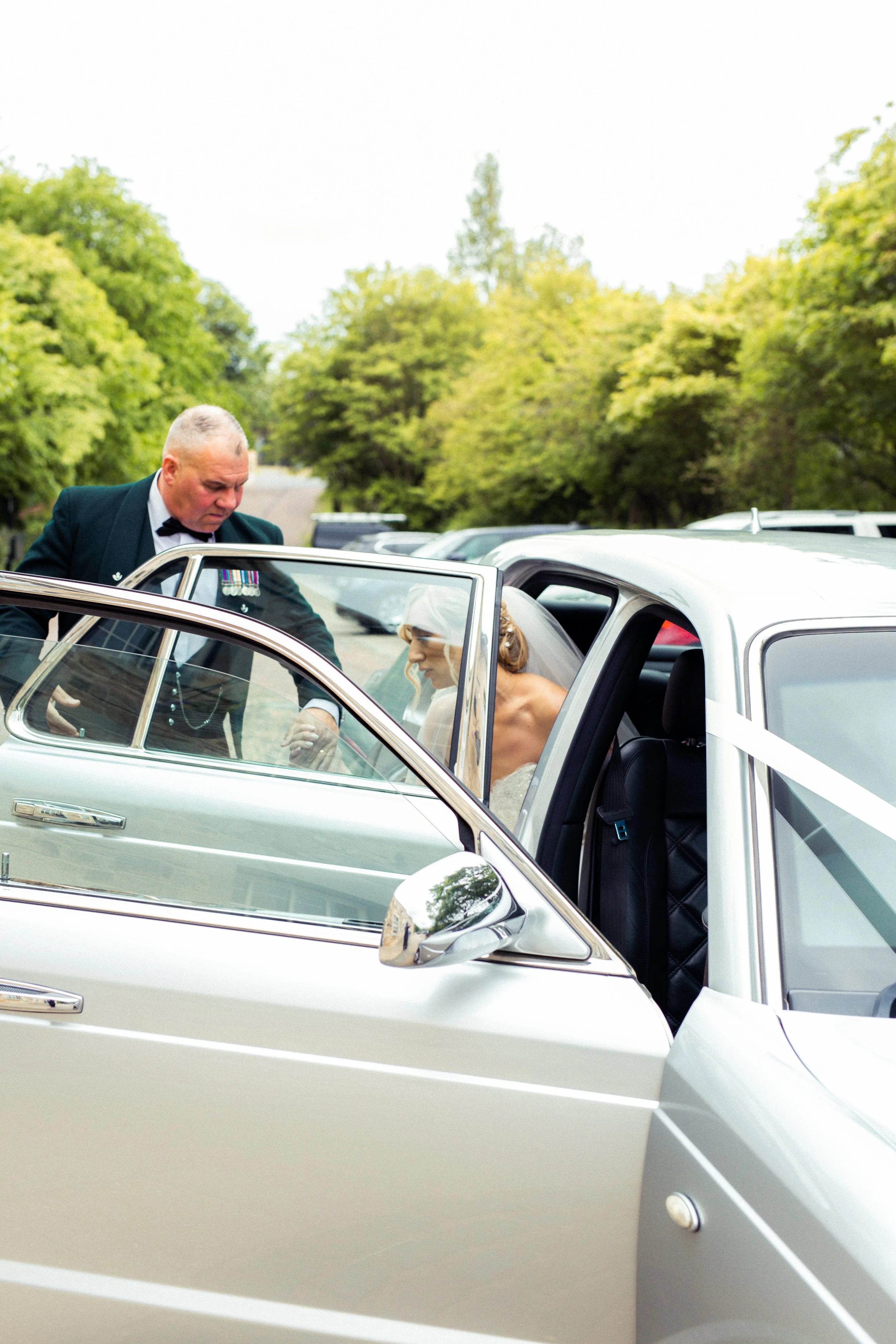 A bride in a white wedding dress and veil sitting in the back seat of a silver car, getting help from a man in a black tuxedo with medals, next to another person. The scene is outdoors with lush green trees in the background.