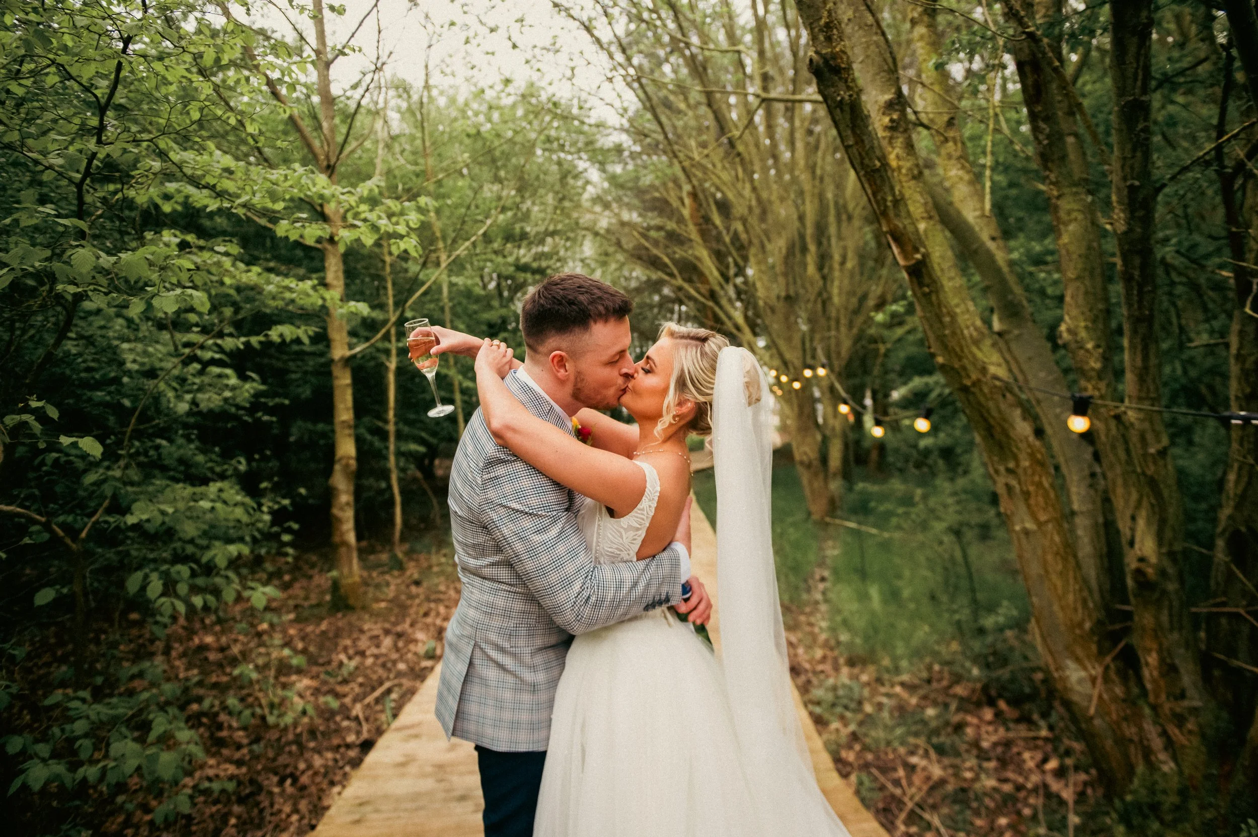 Bride and groom sharing a kiss on a wooded wedding outdoor pathway, with string lights hanging between trees.