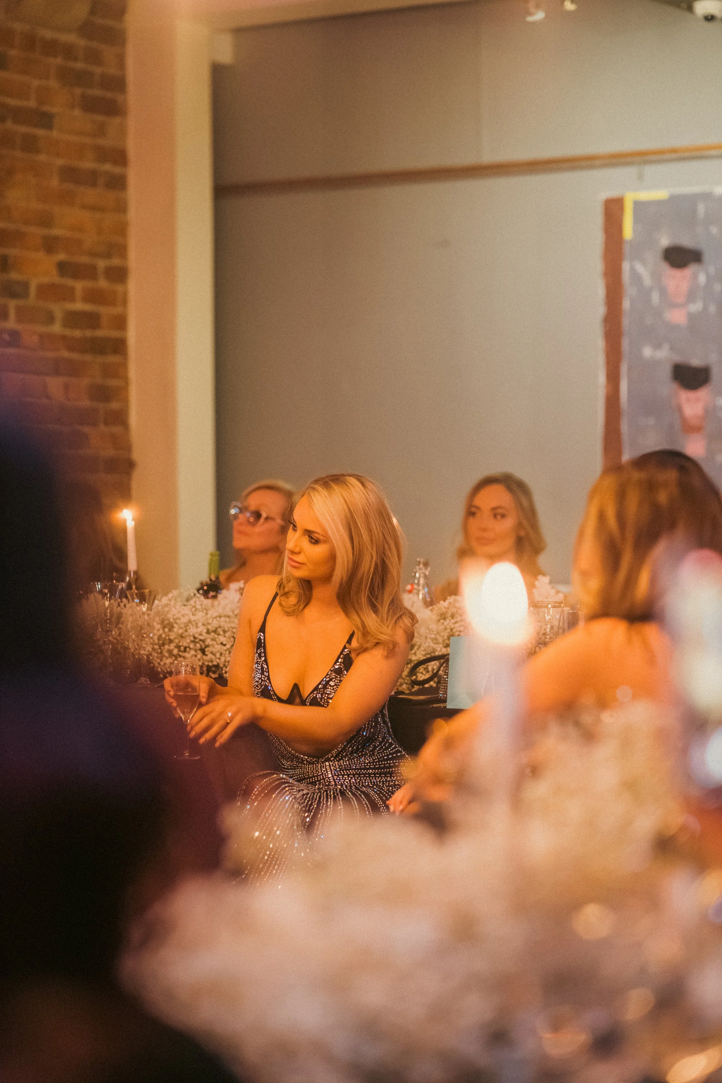 Women sitting at a decorated dinner table during a formal event, surrounded by candles and flowers, in a warmly lit room with brick and gray walls.